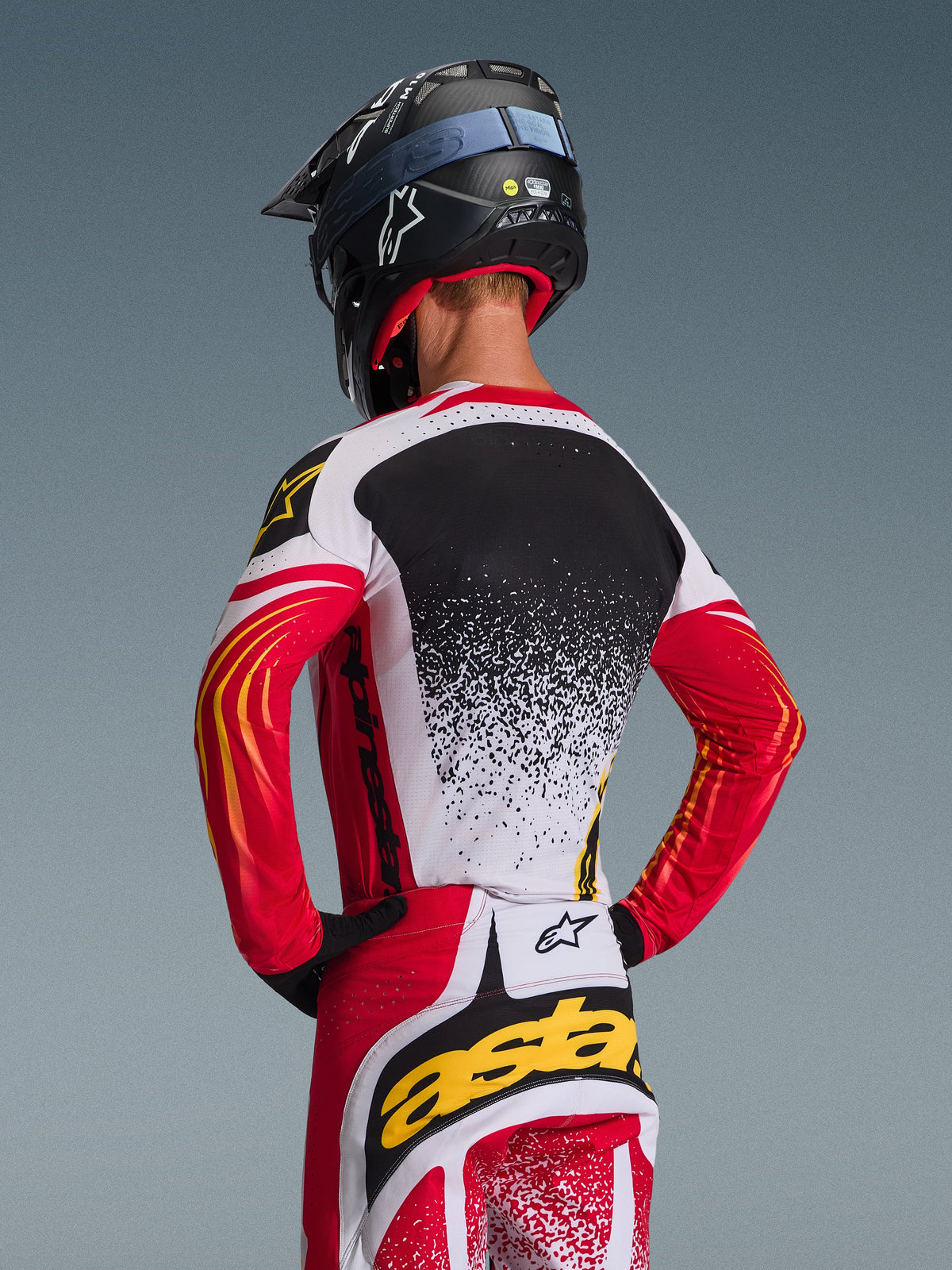 Conjunto de Equipamento Alpinestars Techstar Nomur 2026, camisola e calças de motocross, vermelho, branco e preto com detalhes amarelos, vista traseira de piloto com capacete preto, vestuário de corrida de performance com grafismos de salpicos e logótipos destacados