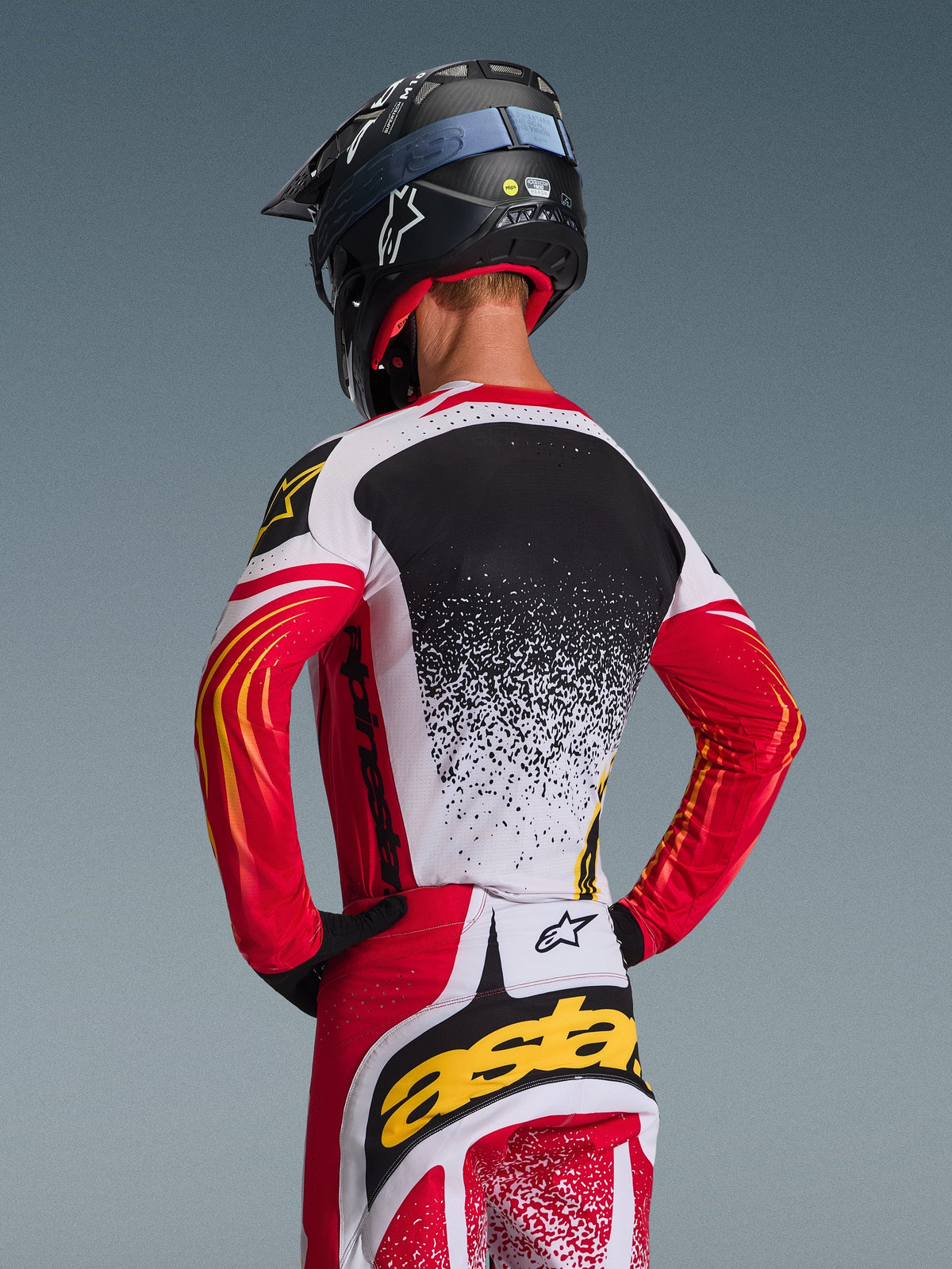 Piloto de motocross visto de costas a usar a camisola Alpinestars 2026 Techstar Nomur e calças a condizer, vermelho, branco e preto com detalhes amarelos, apresentando um design de gradiente salpicado nas costas, capacete Alpinestars preto e luvas pretas, equipamento profissional coordenado para corridas todo-o-terreno
