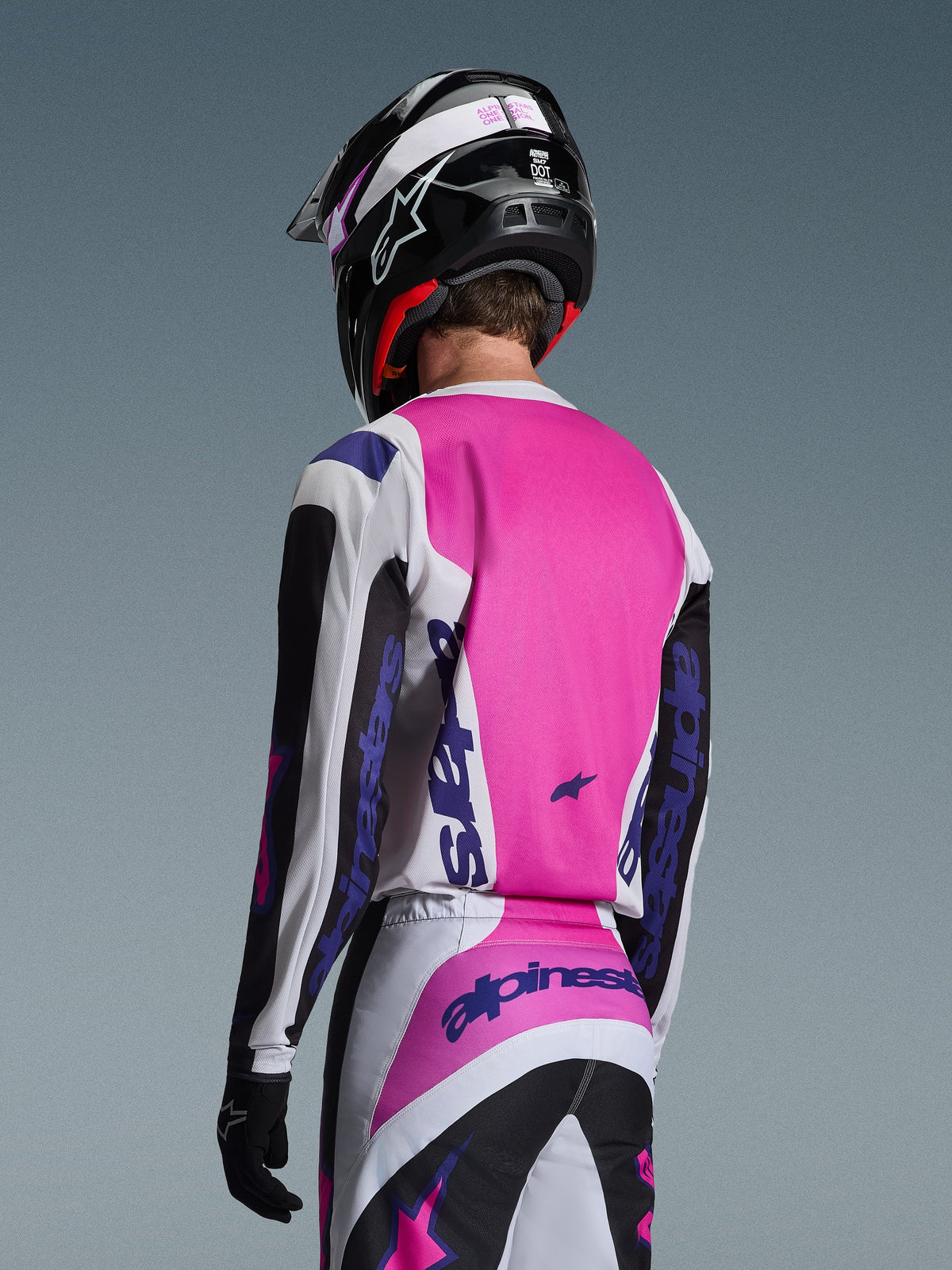 Piloto de motocross visto de costas a usar a camisola Alpinestars 2026 Fluid Grid em preto, branco e roxo, calças a condizer com logótipos destacados, capacete preto com detalhes cor-de-rosa e luvas pretas, equipamento completo coordenado para desempenho todo-o-terreno