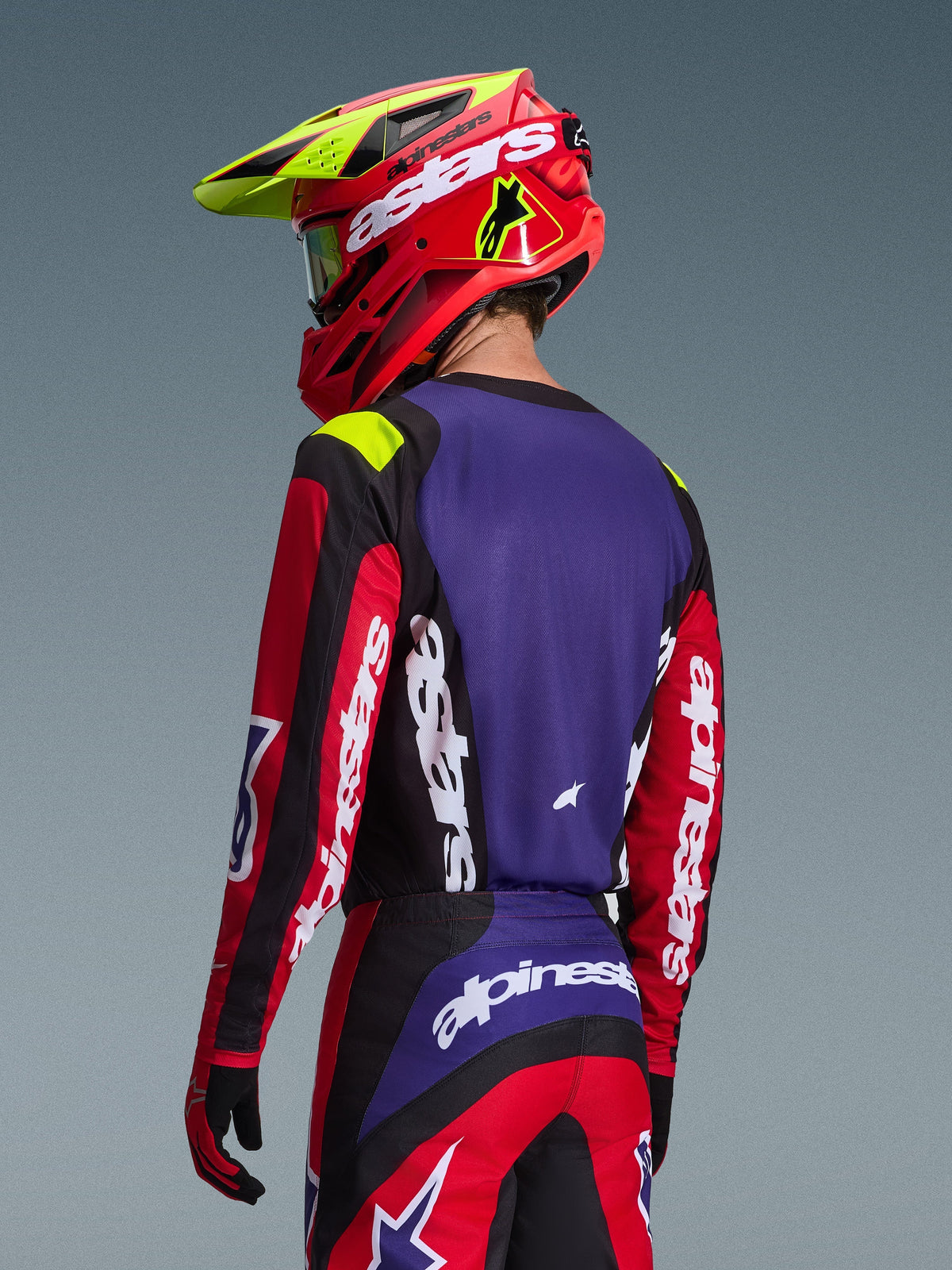 Motocross rider wearing Alpinestars 2026 Fluid Grid Jersey, Red Black and Purple, back view showing purple back panel and red sleeves with white branding, matching pants and a red and neon yellow helmet, coordinated outfit for off-road racing