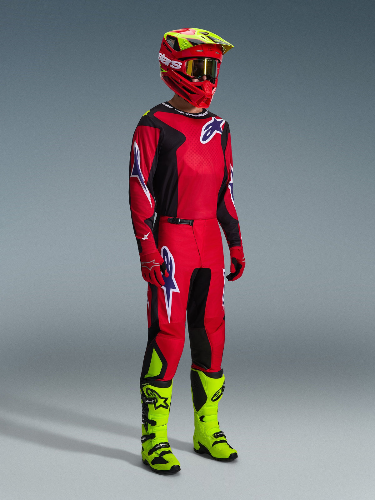 Motocross rider wearing complete racing gear, 2026 Fluid Grid Jersey in Red Black Purple, matching pants with purple logo accents, red and neon yellow helmet with gold goggles, and neon yellow boots, coordinated outfit for off-road racing