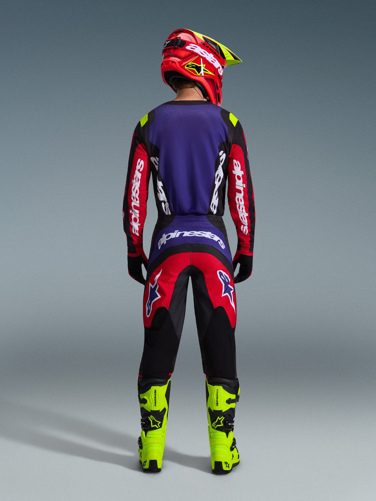 Motocross rider from behind wearing the 2026 Fluid Grid Jersey in Red Black Purple, featuring white Alpinestars branding on long sleeves, paired with matching motocross pants, neon yellow boots, and a red and yellow helmet, complete gear set for off-road racing