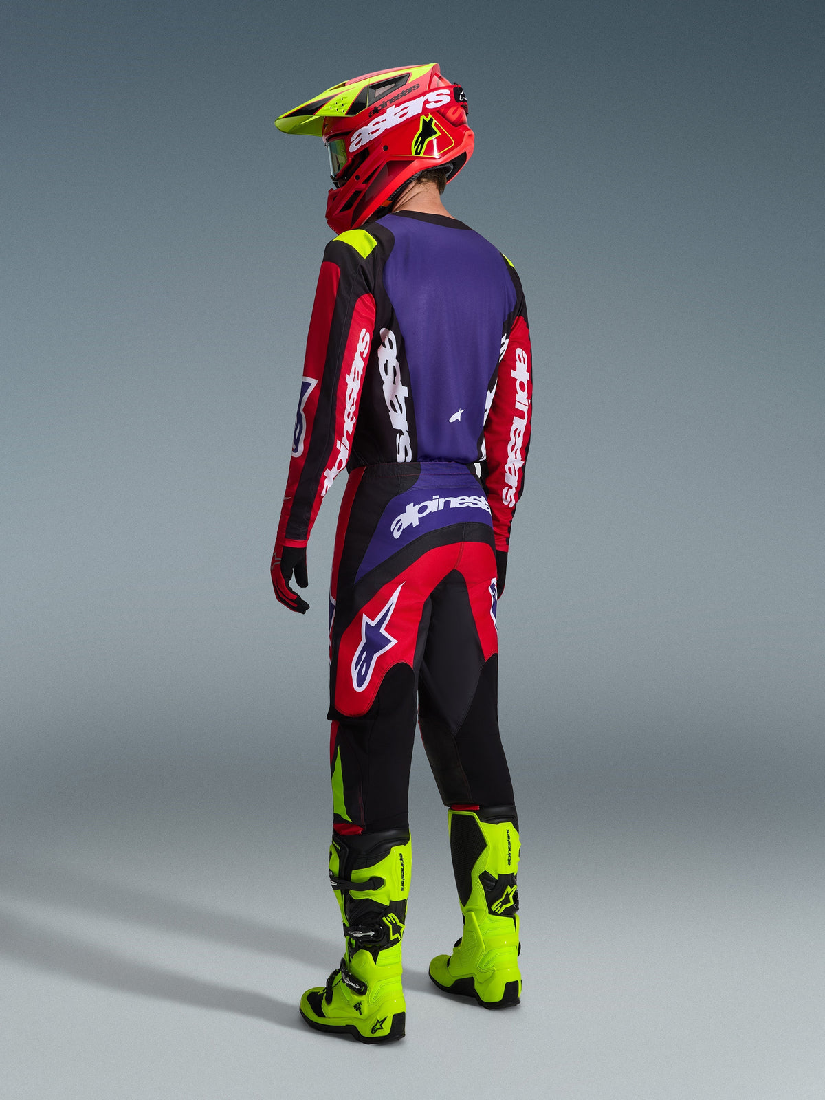Motocross rider seen from the back wearing the Alpinestars 2026 Fluid Grid Jersey and matching pants in Red Black and Purple, accented with white branding, paired with neon yellow boots and a red and yellow helmet, complete off-road racing outfit