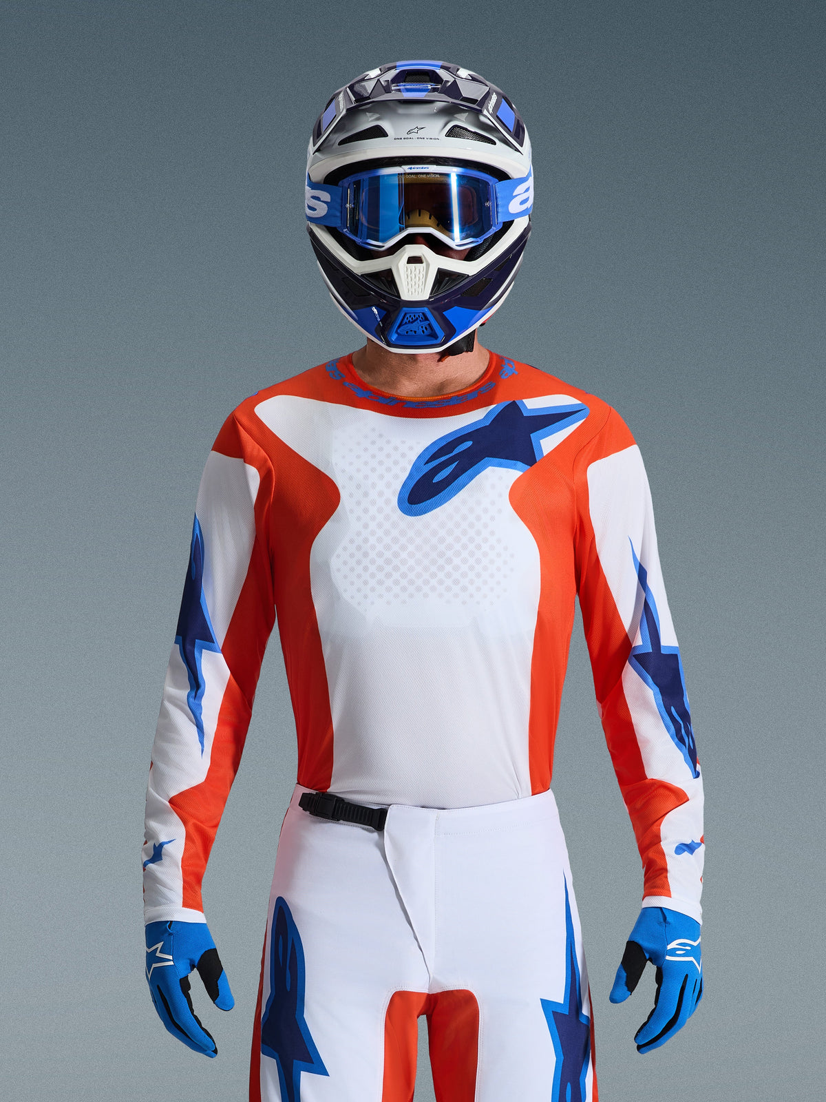 Piloto de motocross com a Camisola Alpinestars Fluid Grid 2026 em Orange Ucla Blue, peito branco com painéis laterais cor de laranja e logótipos azuis, calças brancas e azuis a condizer, luvas azuis e capacete branco e azul, visual profissional de corrida todo-o-terreno