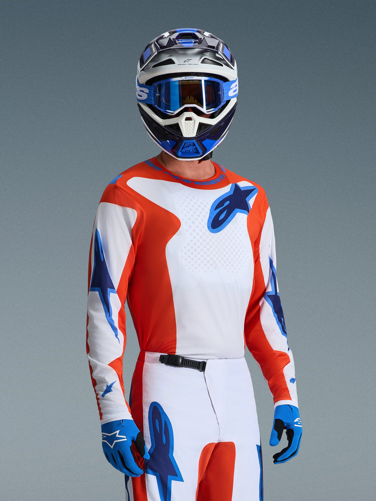 Camisola Alpinestars 2026 Fluid Grid, camisola de motocross em cor de laranja e azul UCLA Blue, usada por um piloto com calças brancas e cor de laranja a condizer, capacete azul e branco, óculos de proteção azuis e luvas azuis, conjunto completo de equipamento de corrida todo-o-terreno