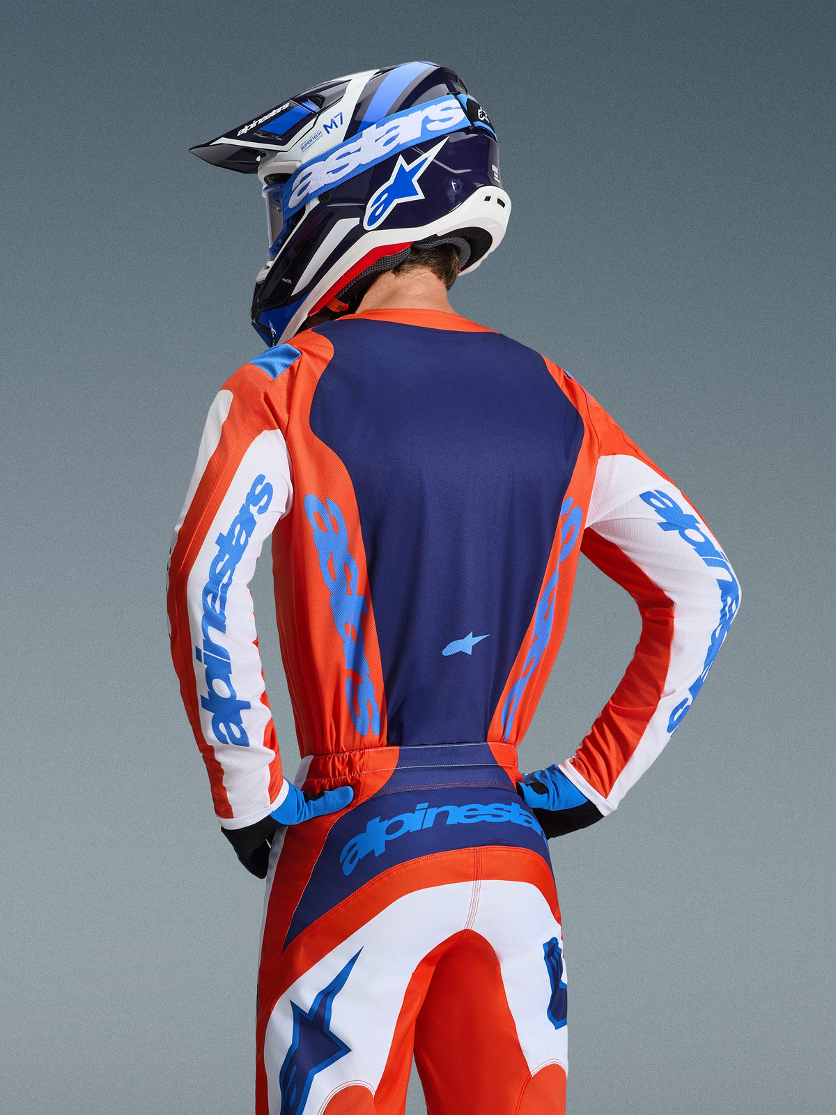 Alpinestars 2026 Fluid Grid Jersey, camisola de motocross, laranja e azul UCLA, vista traseira do piloto a usar calças e capacete a condizer, com painel traseiro azul-marinho e mangas brancas com logótipo azul-claro, corte atlético para corridas de todo-o-terreno