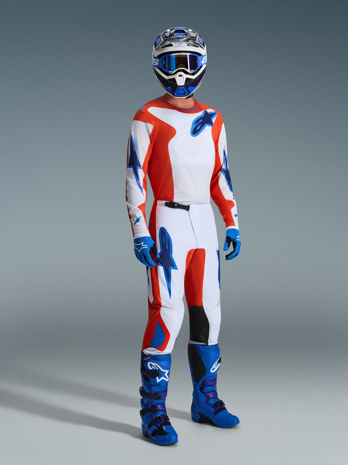Piloto de motocross com equipamento de corrida completo, Camisola Fluid Grid 2026 em cor de laranja e azul UCLA, calças a condizer com painéis brancos e logótipos azuis, botas de motocross azuis e capacete coordenado, fotografia de estúdio profissional para corridas todo-o-terreno.