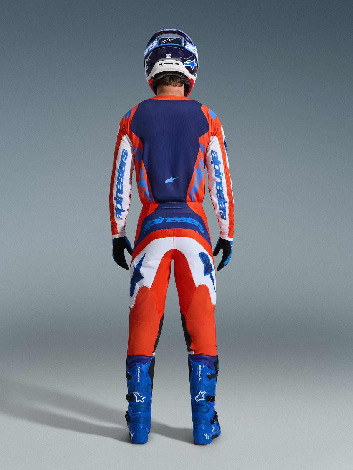 Alpinestars 2026 Fluid Grid Jersey, camisola de motocross, laranja e azul UCLA, vista traseira do piloto com o conjunto completo de equipamento de corrida, com um painel traseiro azul escuro e mangas brancas com logótipo azul, combinada com calças a condizer e botas azuis.
