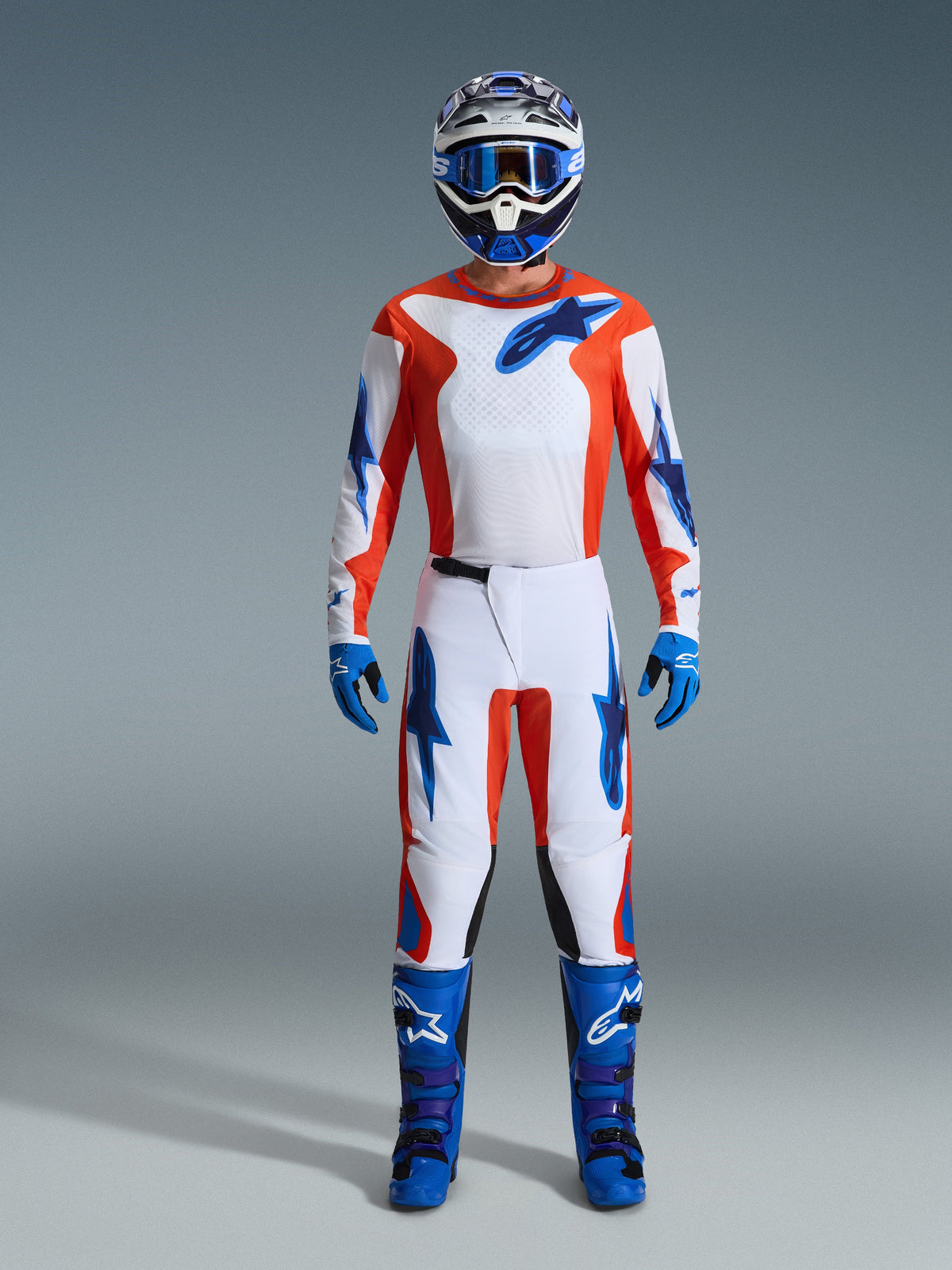 Piloto de motocross com equipamento completo de competição, camisola Fluid Grid 2026 em laranja e azul UCLA, calças brancas a condizer com detalhes em laranja e azul, capacete azul e branco com óculos azuis, botas de todo-o-terreno azuis e roxas, luvas azuis, conjunto coordenado para corridas de motocross.