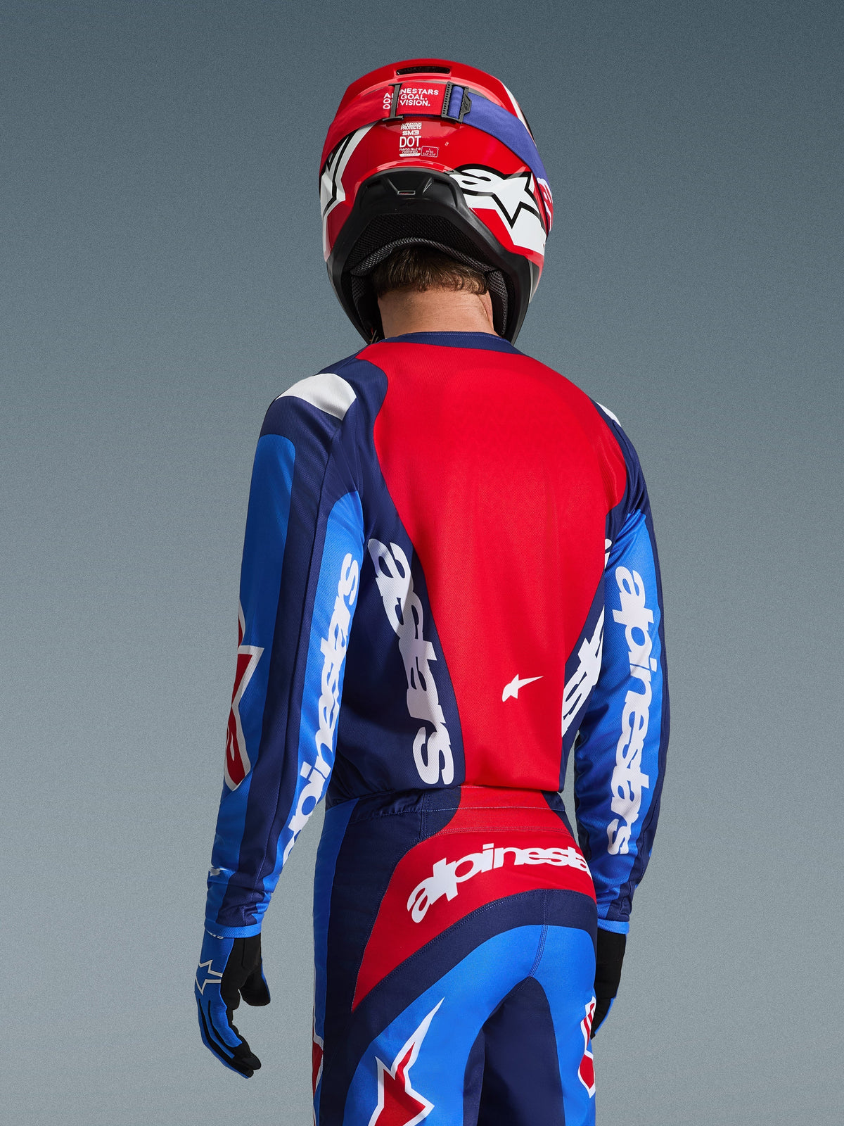 Vista traseira de um piloto de motocross a usar a camisola Alpinestars 2026 Fluid Grid em azul e vermelho, com logótipos brancos destacados nas mangas e nas costas, combinada com calças de motocross a condizer e um capacete vermelho para um visual completo de corrida todo-o-terreno