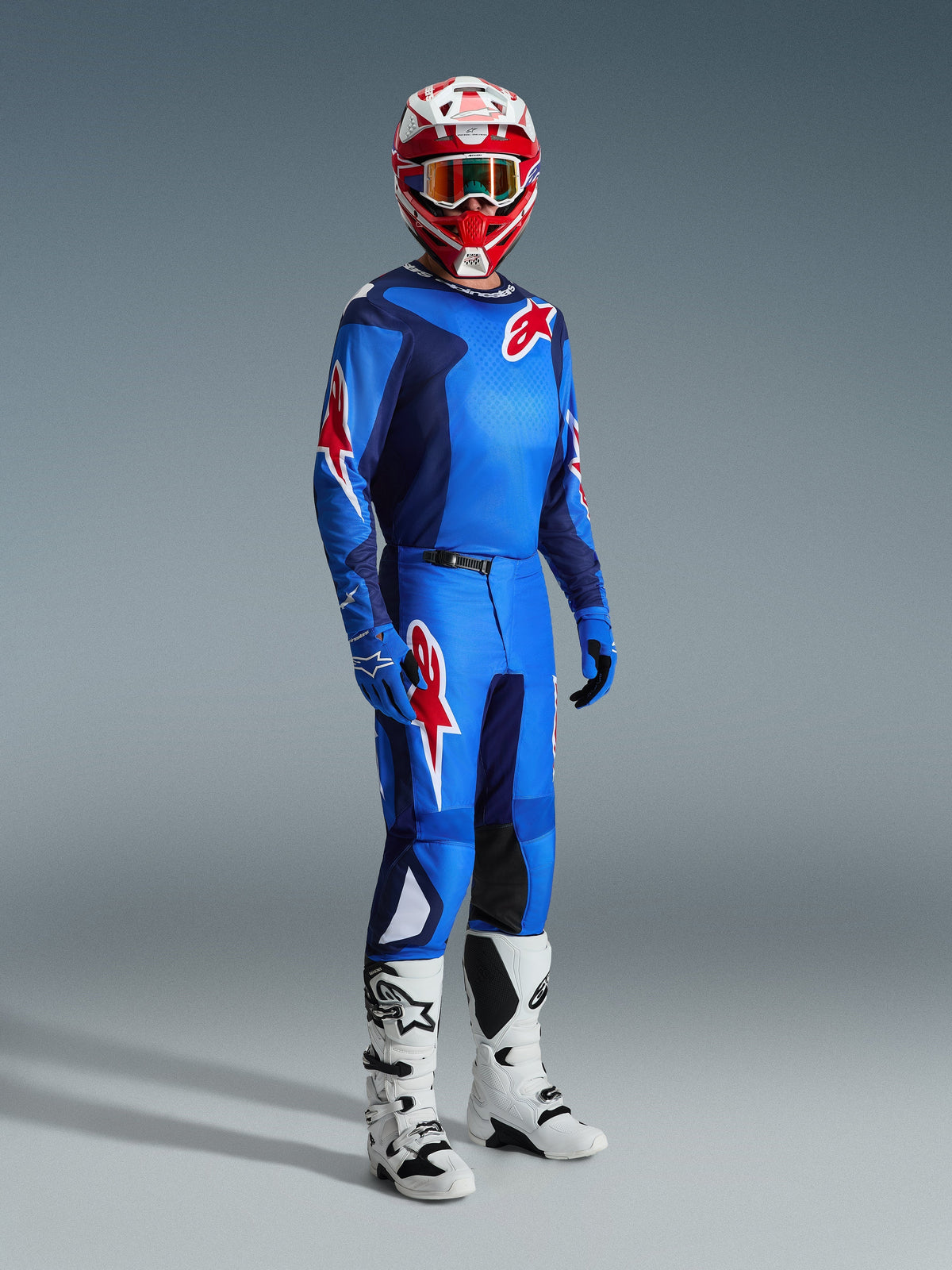 Piloto de motocross a usar a camisola Alpinestars 2026 Fluid Grid em Azul e Vermelho, acompanhada por calças de motocross a condizer, botas brancas e pretas e capacete vermelho e branco, conjunto de equipamento profissional para desempenho todo-o-terreno.