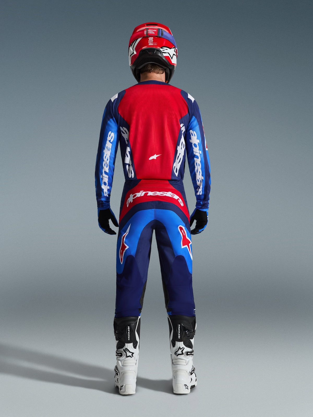 Vista traseira de um piloto de motocross a usar a Camisola Fluid Grid 2026 em Azul e Vermelho, com um grande painel traseiro vermelho e mangas azuis com a marca Alpinestars a branco, combinada com calças a condizer, botas brancas e pretas, e um capacete vermelho para um equipamento completo de corrida todo-o-terreno.