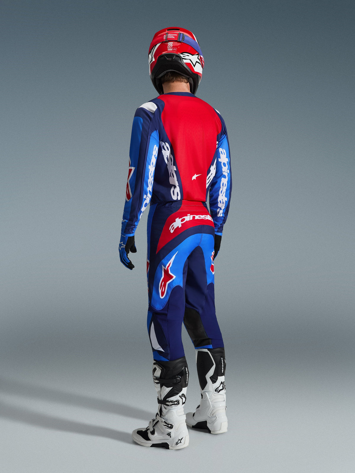 Piloto de motocross visto de costas a usar a camisola Alpinestars 2026 Fluid Grid em azul e vermelho, calças de motocross a combinar com branding branco destacado, capacete vermelho e botas brancas e pretas, conjunto completo de equipamento de corrida off-road