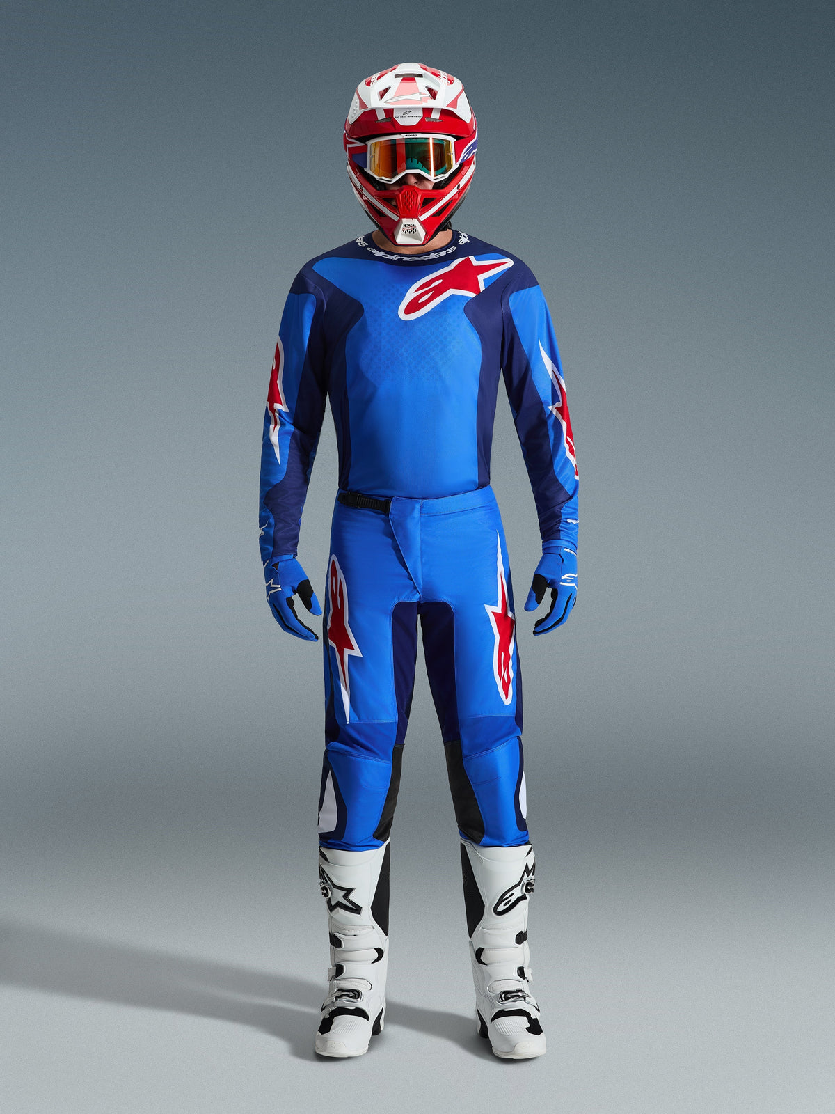 Piloto de motocross a usar a camisola Alpinestars 2026 Fluid Grid em azul e vermelho, calças azuis a condizer com logótipos de estrela em branco e vermelho, capacete vermelho e branco com óculos espelhados, luvas azuis e botas de competição brancas e pretas, conjunto completo de equipamento para todo-o-terreno