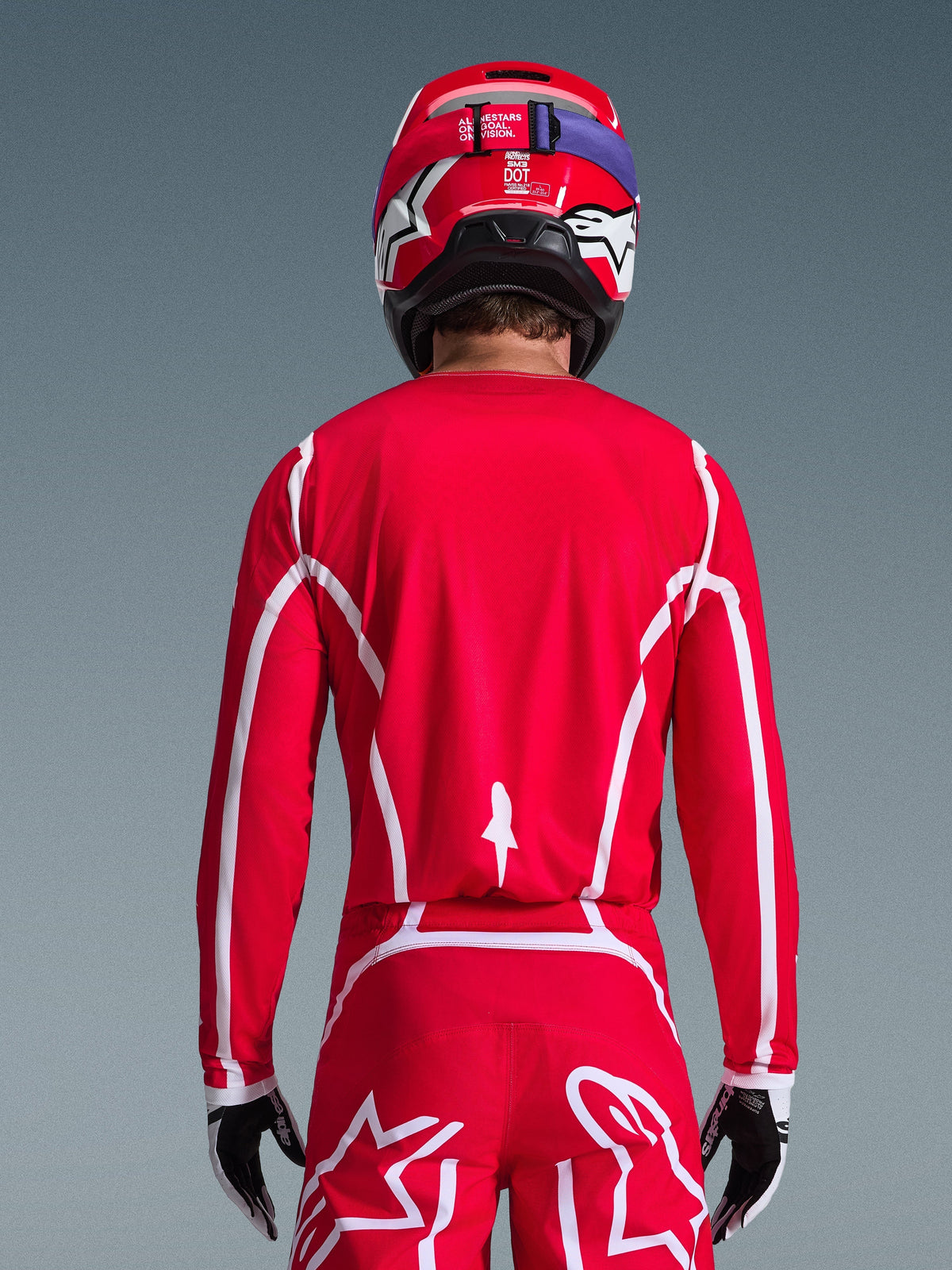 Vista traseira de um piloto de motocross a usar a camisola Alpinestars 2026 Fluid Apex em vermelho e branco, com padrões geométricos brancos, combinada com calças de motocross a condizer e um capacete vermelho, equipamento de corrida completo para desempenho off-road.