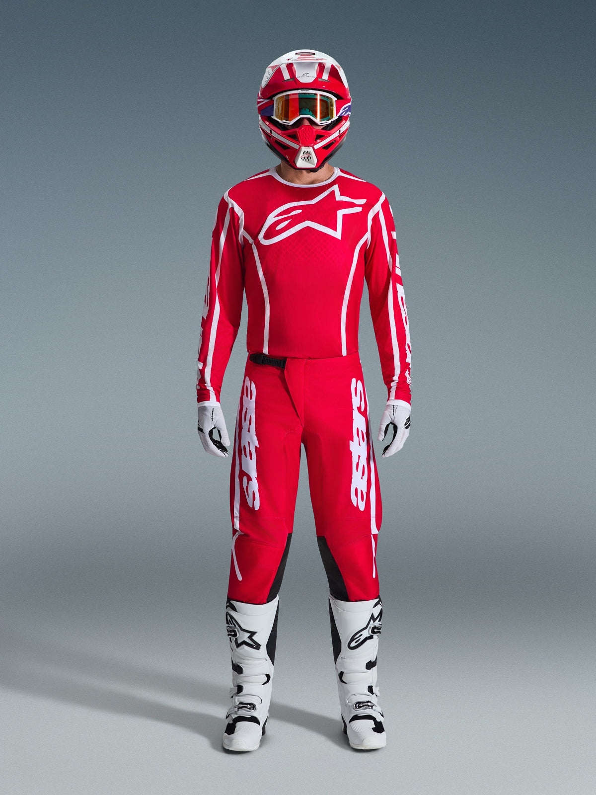 Piloto de motocross com o conjunto completo de equipamento Alpinestars 2026 Fluid Apex, camisola vermelha com logótipo e marca nas mangas em branco, calças vermelhas com detalhes brancos e painéis pretos, capacete vermelho e branco com óculos espelhados, botas brancas e pretas e luvas brancas e pretas, conjunto de alta performance para off-road