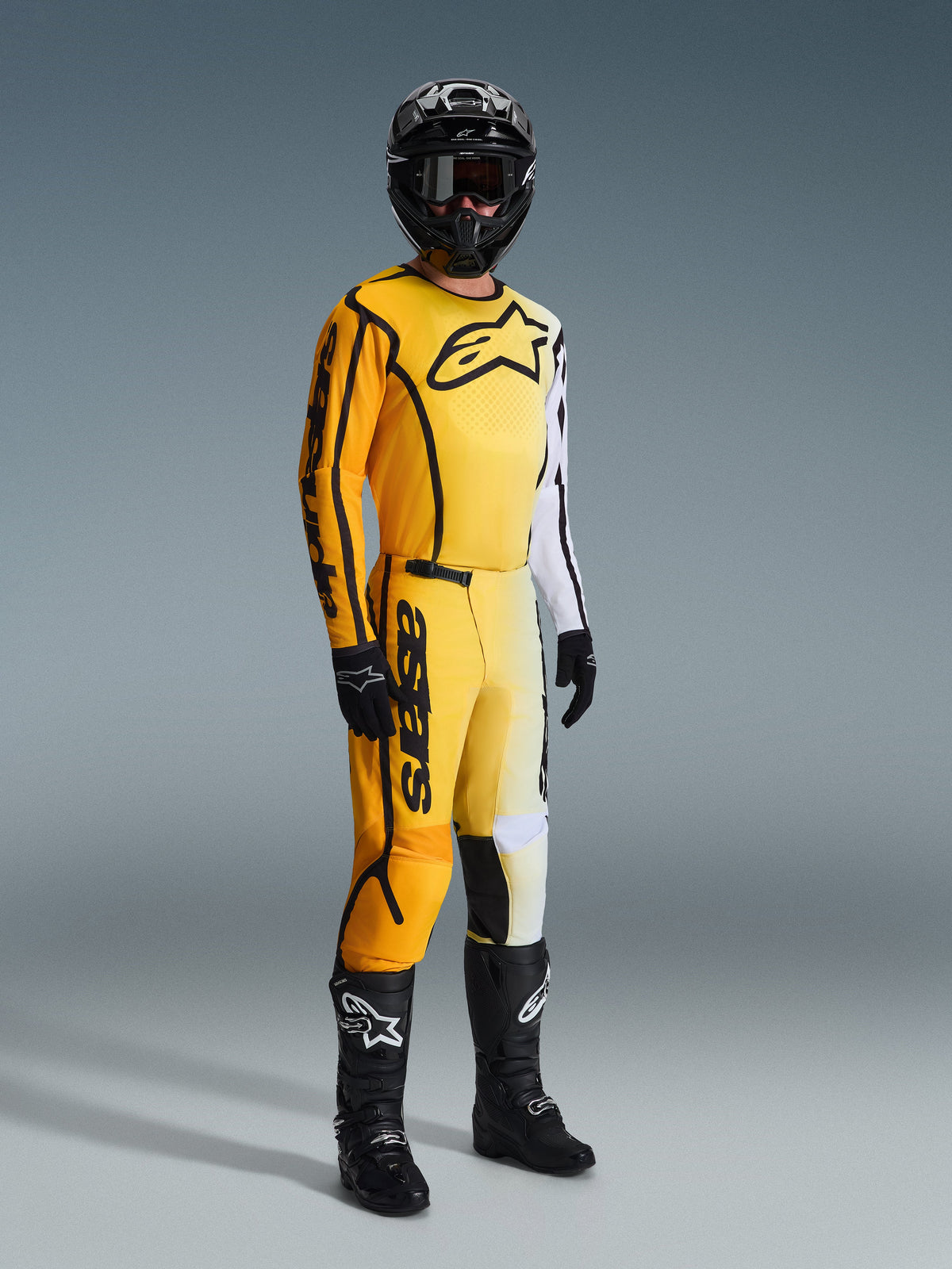 Conjunto de Equipamento Alpinestars 2026 Fluid Apex, piloto de motocross com equipamento de corrida amarelo, branco e preto, camisola com logótipo grande no peito e manga branca, calças a condizer com marca em destaque, capacete, luvas e botas pretos, conjunto coordenado para corridas todo-o-terreno
