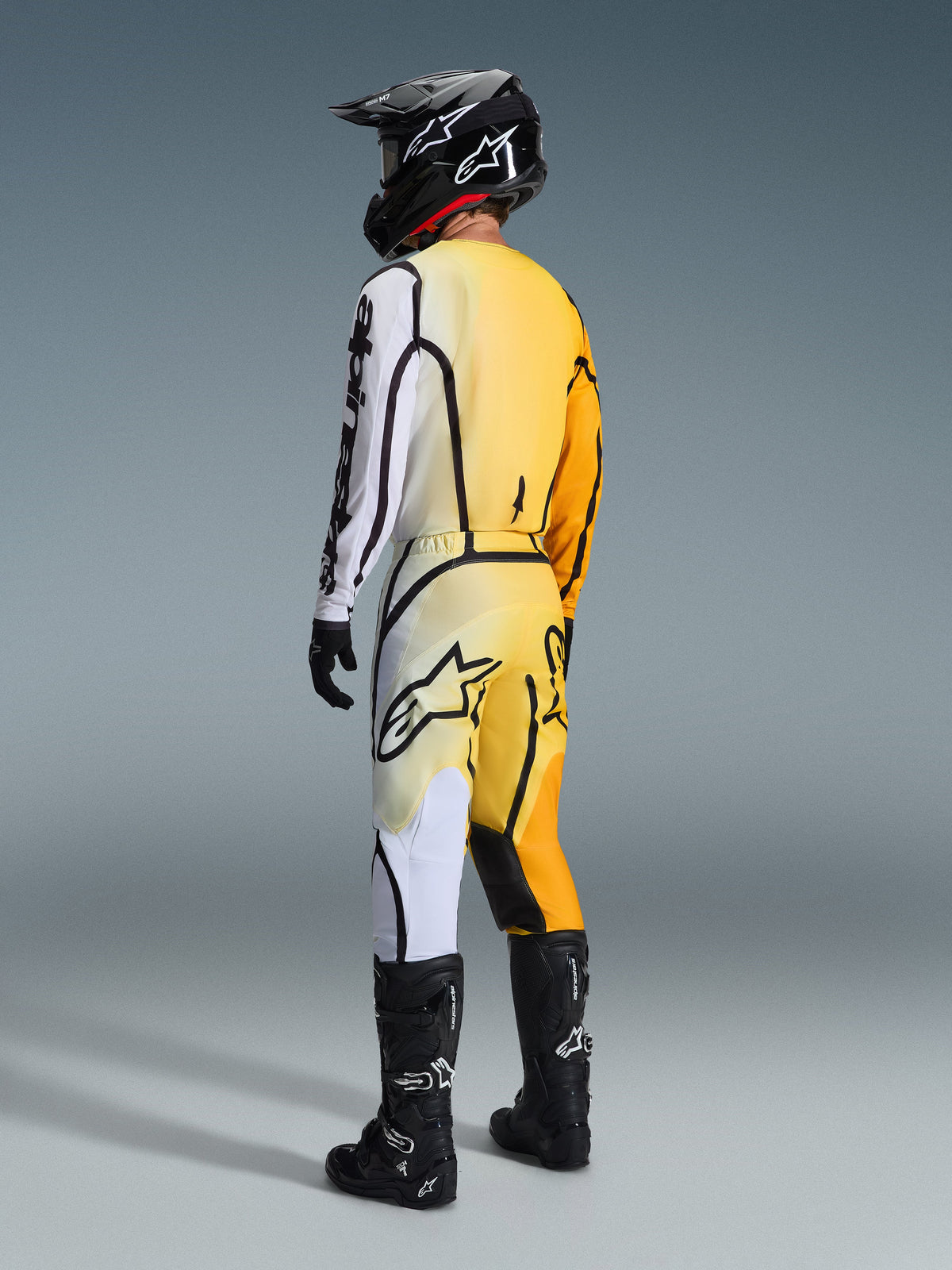 Motocross rider seen from behind wearing the Alpinestars 2026 Fluid Apex Jersey and matching pants, Yellow Black gear set with yellow-to-white gradients and bold black graphic lines, paired with a black helmet, black and white gloves, and black motocross boots