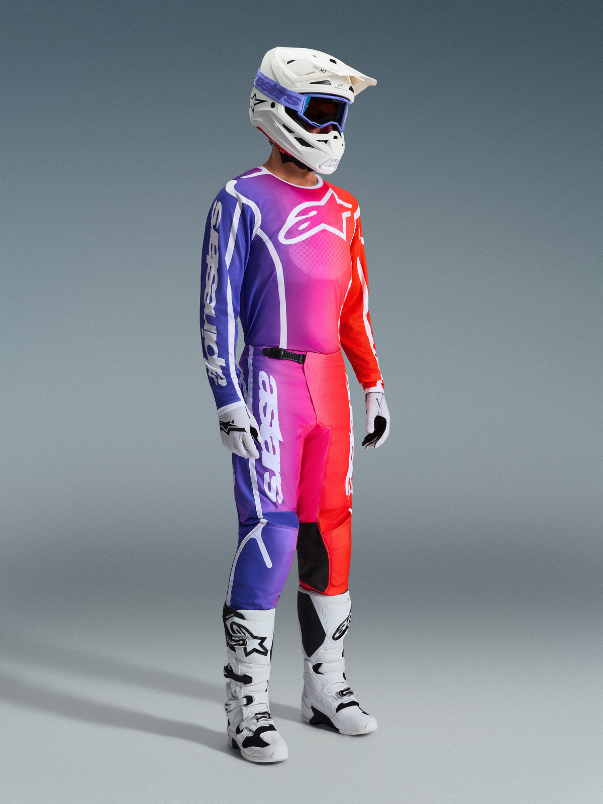 Alpinestars 2026 Fluid Apex Gear Set, piloto de motocross com um conjunto de corrida completo em degradê vibrante de roxo, rosa e vermelho, capacete branco com óculos roxos, botas brancas e pretas, luvas brancas, visual atlético coordenado para desempenho off-road