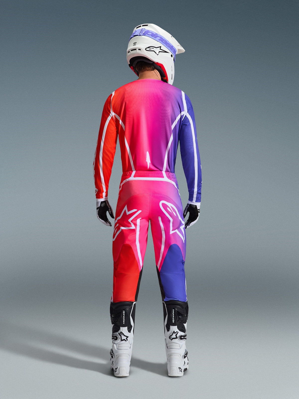 Conjunto de Equipamento Alpinestars Fluid Apex 2026, vista traseira de um piloto de motocross a usar camisola e calças com gradiente vibrante de vermelho, rosa e roxo, linhas gráficas brancas e grandes logótipos de estrela, complementado por capacete e botas brancas.