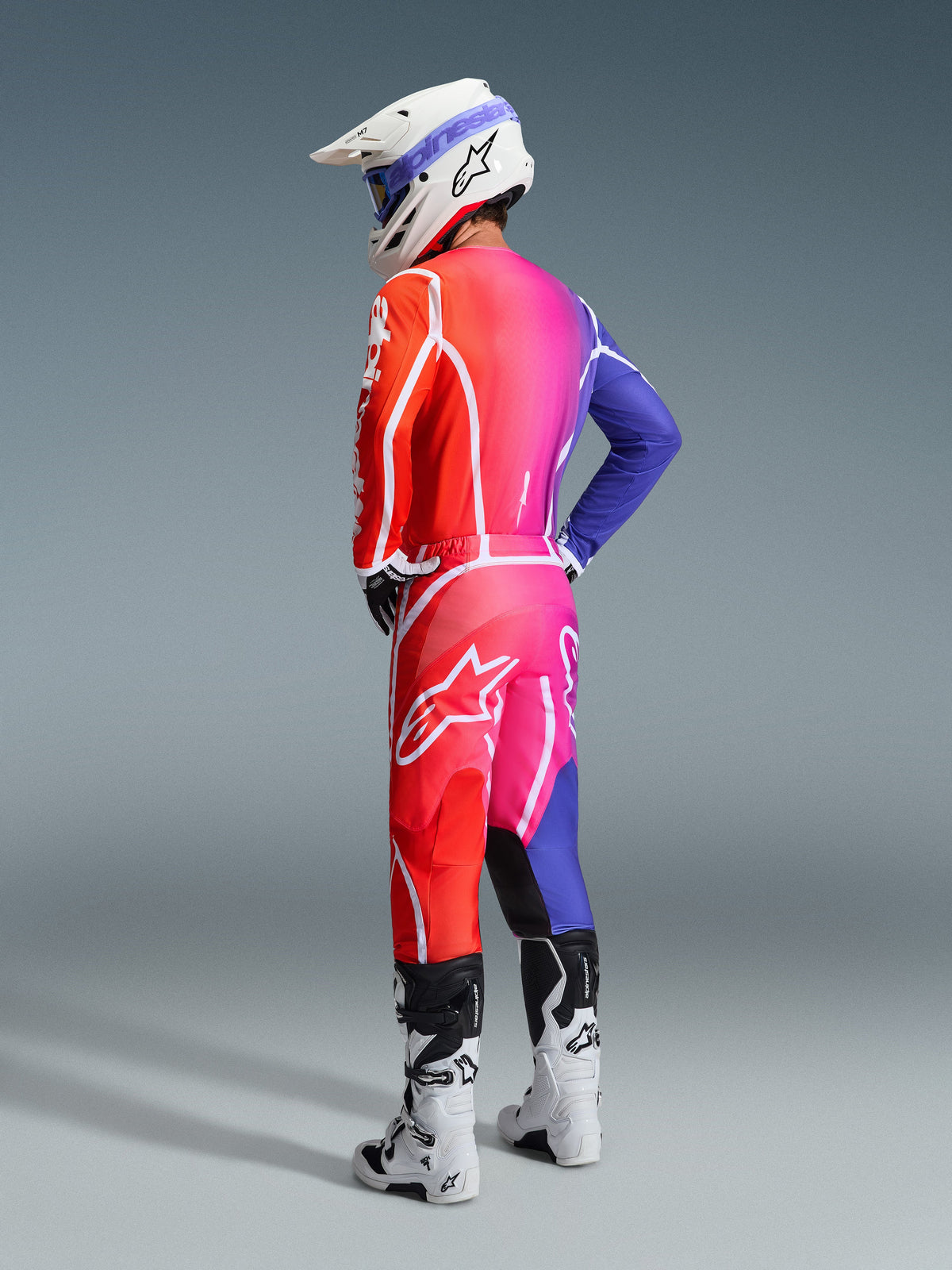 Piloto de motocross com o conjunto de equipamento Alpinestars 2026 Fluid Apex, transição vibrante em degradê de laranja para rosa e roxo com branding branco marcante e logótipos de estrela, combinado com capacete branco, óculos azuis e botas brancas, visto de um ângulo de três quartos traseiro em estúdio