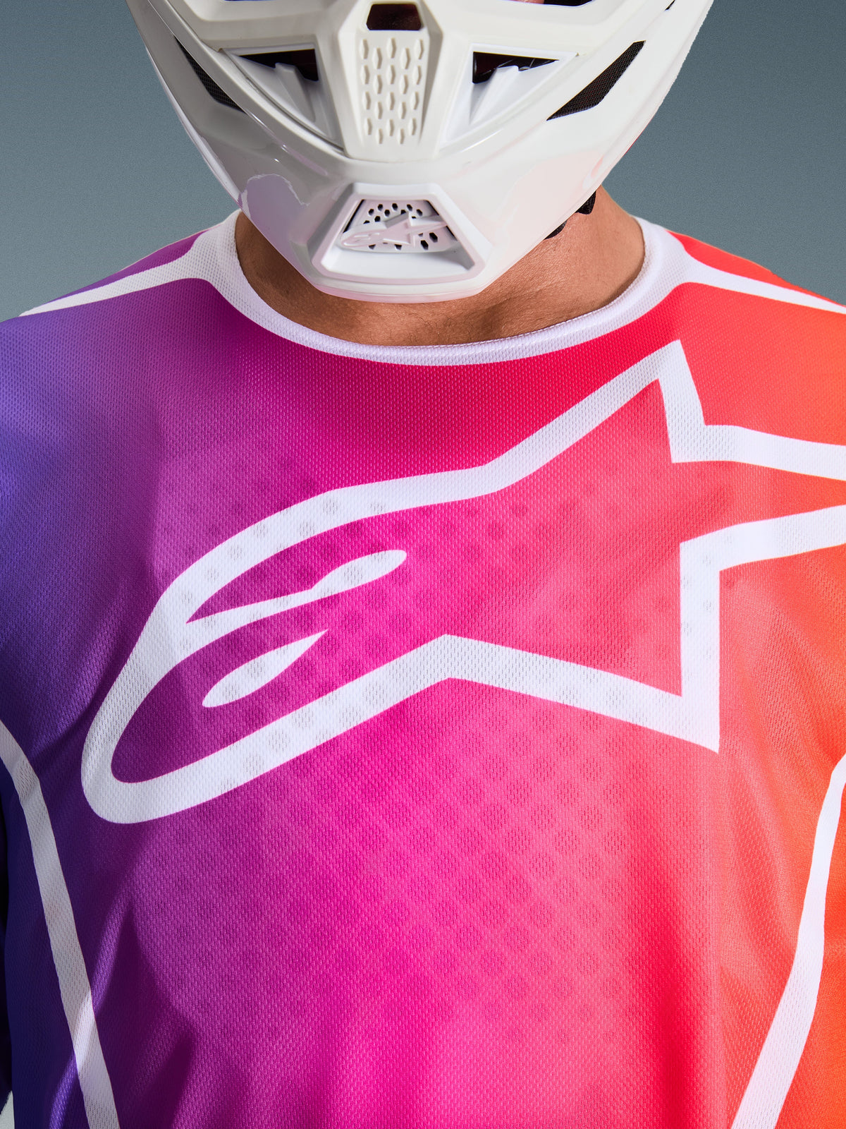 Camisola Alpinestars Fluid Apex 2026, camisola de motocross, degradê roxo, rosa e laranja, grande logótipo de estrela Alpinestars branco no peito, vista de perto mostrando a queixeira de um capacete branco
