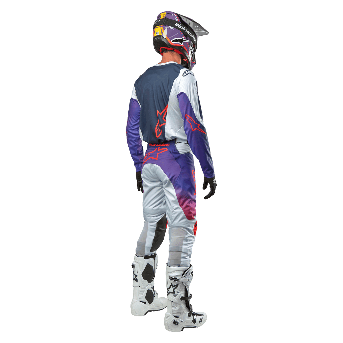 Piloto de motocross visto de costas a usar a camisola Alpinestars 2024 Racer Hoen em cinzento claro, laranja vibrante e preto, painel traseiro azul-marinho com logótipo de estrela cor de laranja, mangas roxas, acompanhada por calças de motocross a condizer e botas brancas.