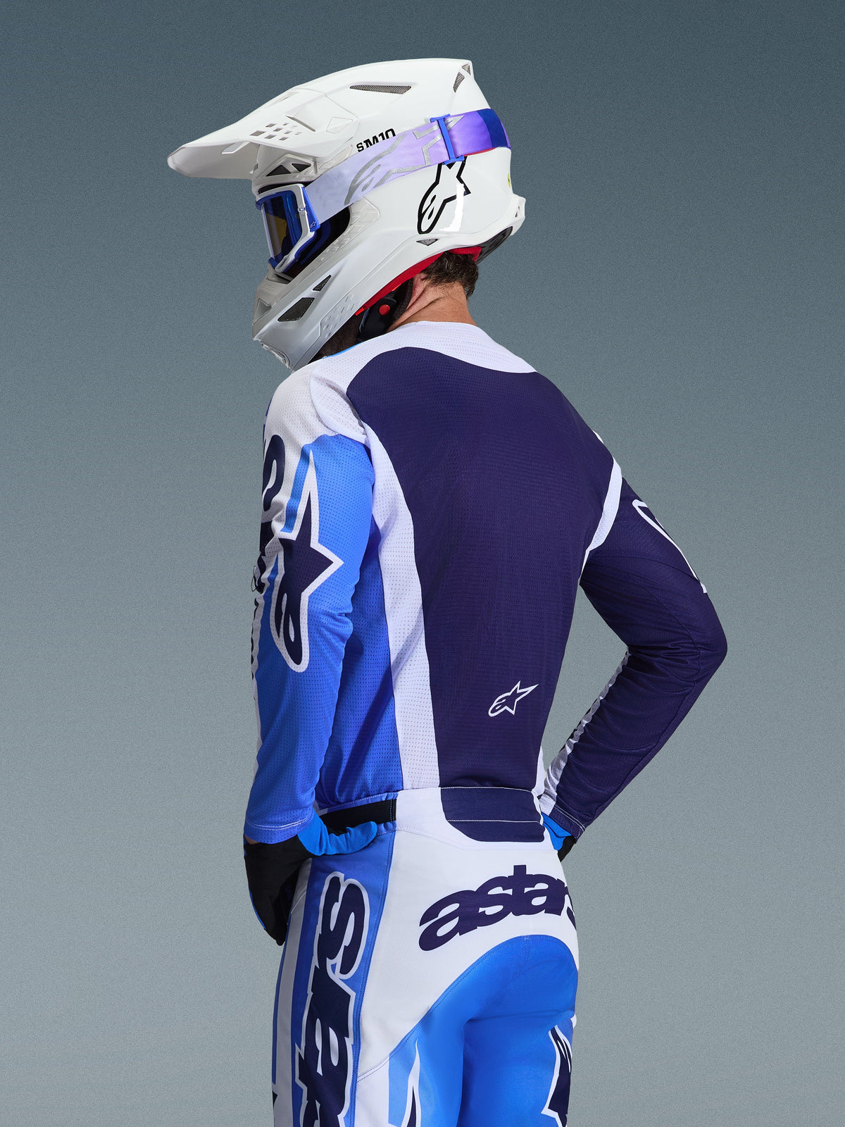 Piloto de motocross com camisola Alpinestars 2026 Racer Air Portl em azul Ucla e branco, vista traseira destacando o painel de malha respirável azul marinho, usada com calças a condizer e capacete Alpinestars branco com óculos azuis, conjunto de equipamento de motocross coordenado