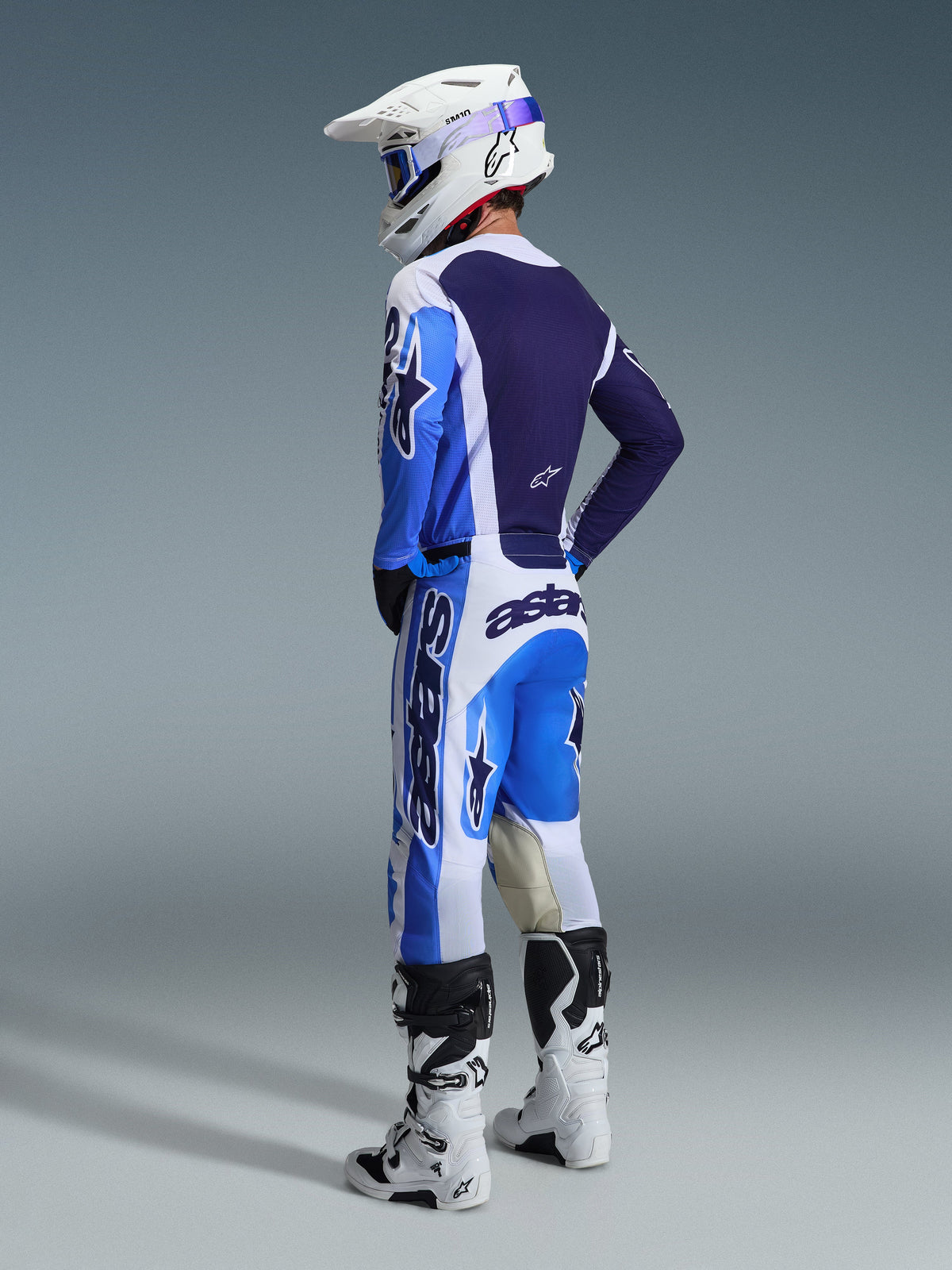 Piloto de motocross visto de uma perspetiva traseira com a camisola Alpinestars 2026 Racer Air Portl em Azul Ucla e Branco, calças de motocross a condizer, capacete Supertech branco e botas brancas, exibindo um conjunto de equipamento de corrida completo e coordenado.