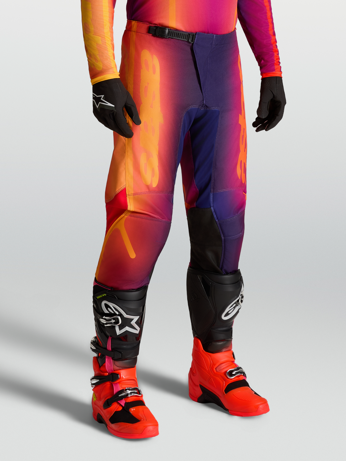 Alpinestars Calças Fluid Kepler, calças de motocross todo-o-terreno roxas e cor de laranja, ajuste desportivo com joelhos reforçados, usadas com botas e luvas pretas.
