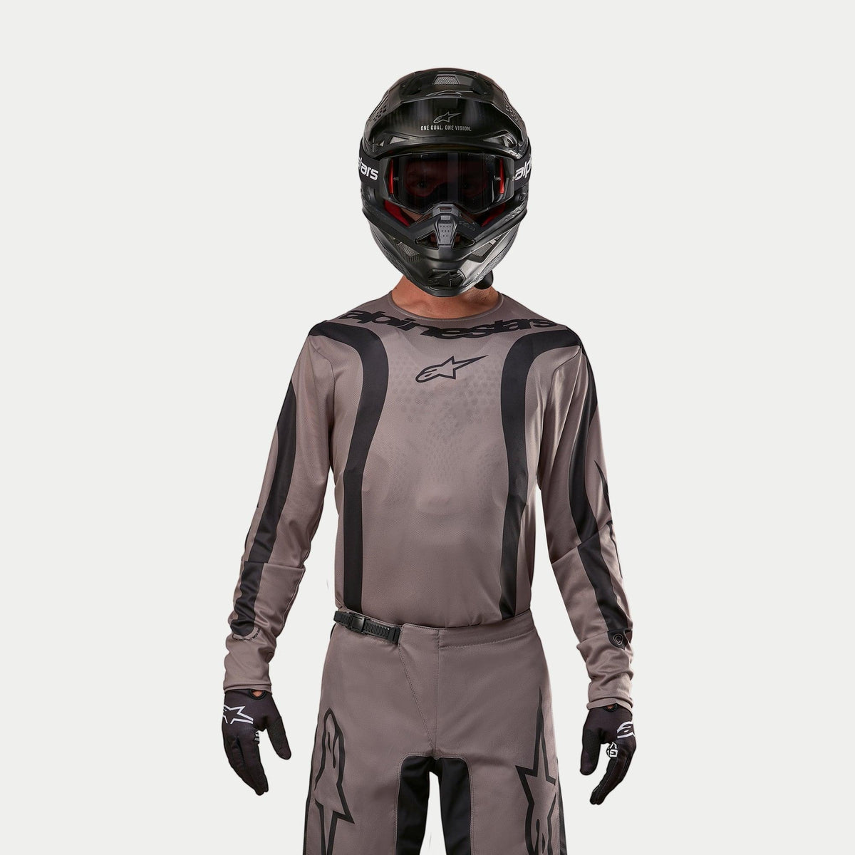 Piloto de motocross a usar a Camisola Alpinestars Fluid Lurv 2024 em Mud Black, um tecido cinzento-castanho terra com riscas gráficas pretas e um logótipo de estrela no peito, combinada com um capacete preto, óculos de proteção vermelhos e luvas pretas para um visual de corrida todo-o-terreno coordenado.