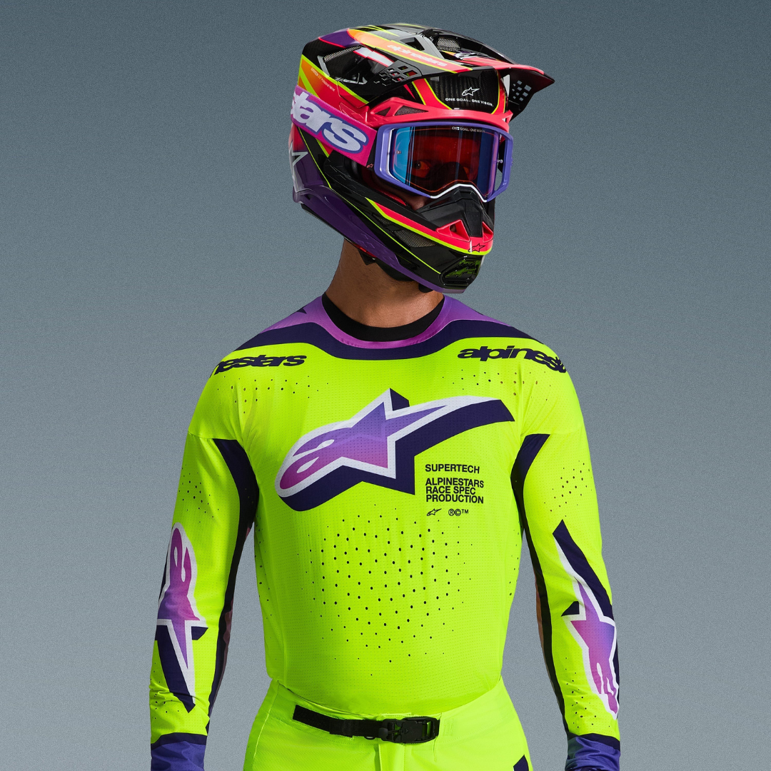 Motocross rider wearing Alpinestars 2026 Supertech Vista Gear, neon yellow jersey with purple and black details, large purple gradient logo on chest, matching vibrant multi-color helmet and purple goggles, showcasing laser-cut ventilation and professional athletic fit