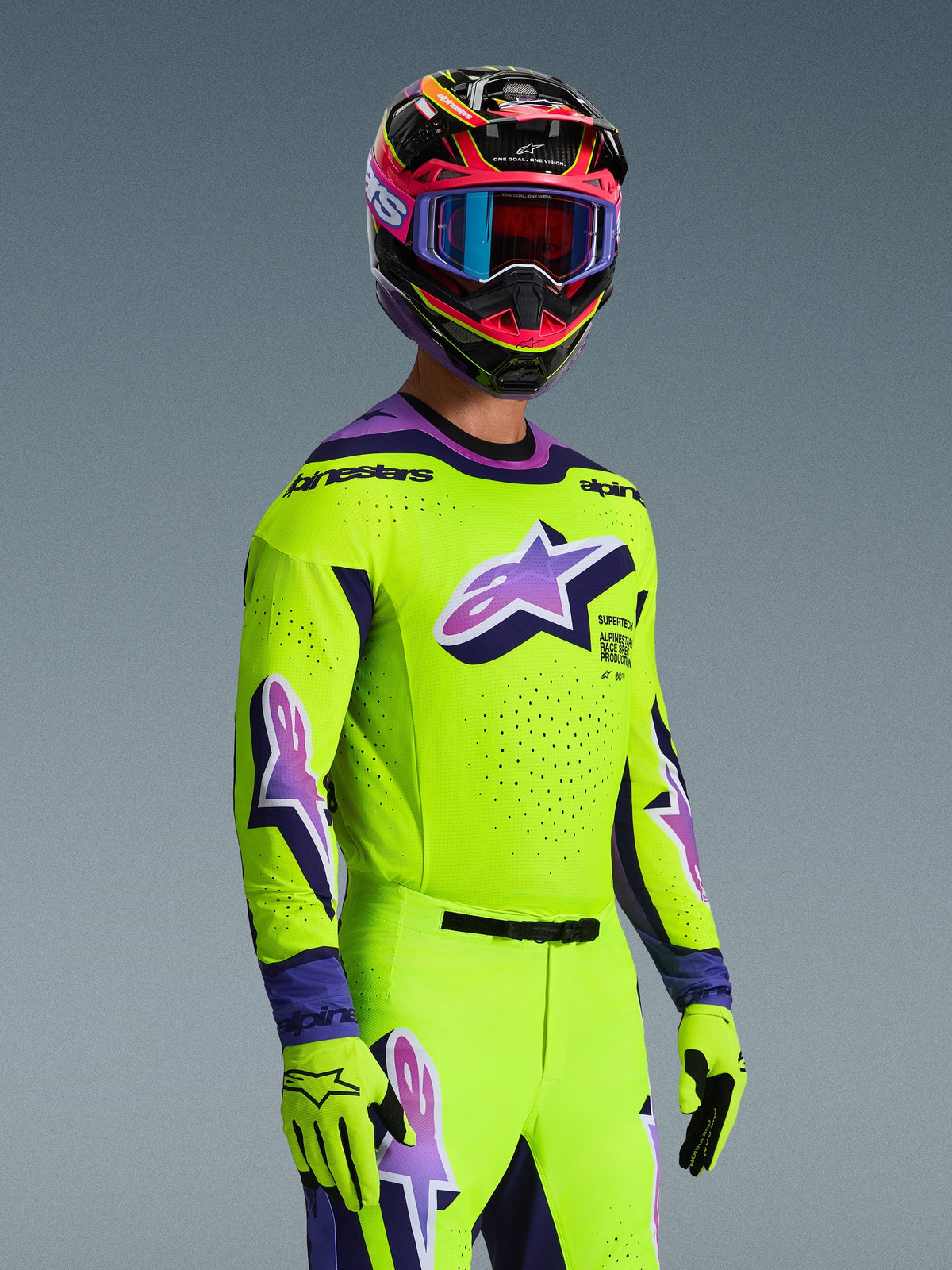 Motocross rider wearing the Alpinestars 2026 Supertech Vista Jersey and matching gear set, bright yellow with purple and black accents, featuring a large purple Astar logo on the chest and sleeves, matching gloves, and a multi-colored racing helmet with goggles, pro-level athletic fit for off-road racing.
