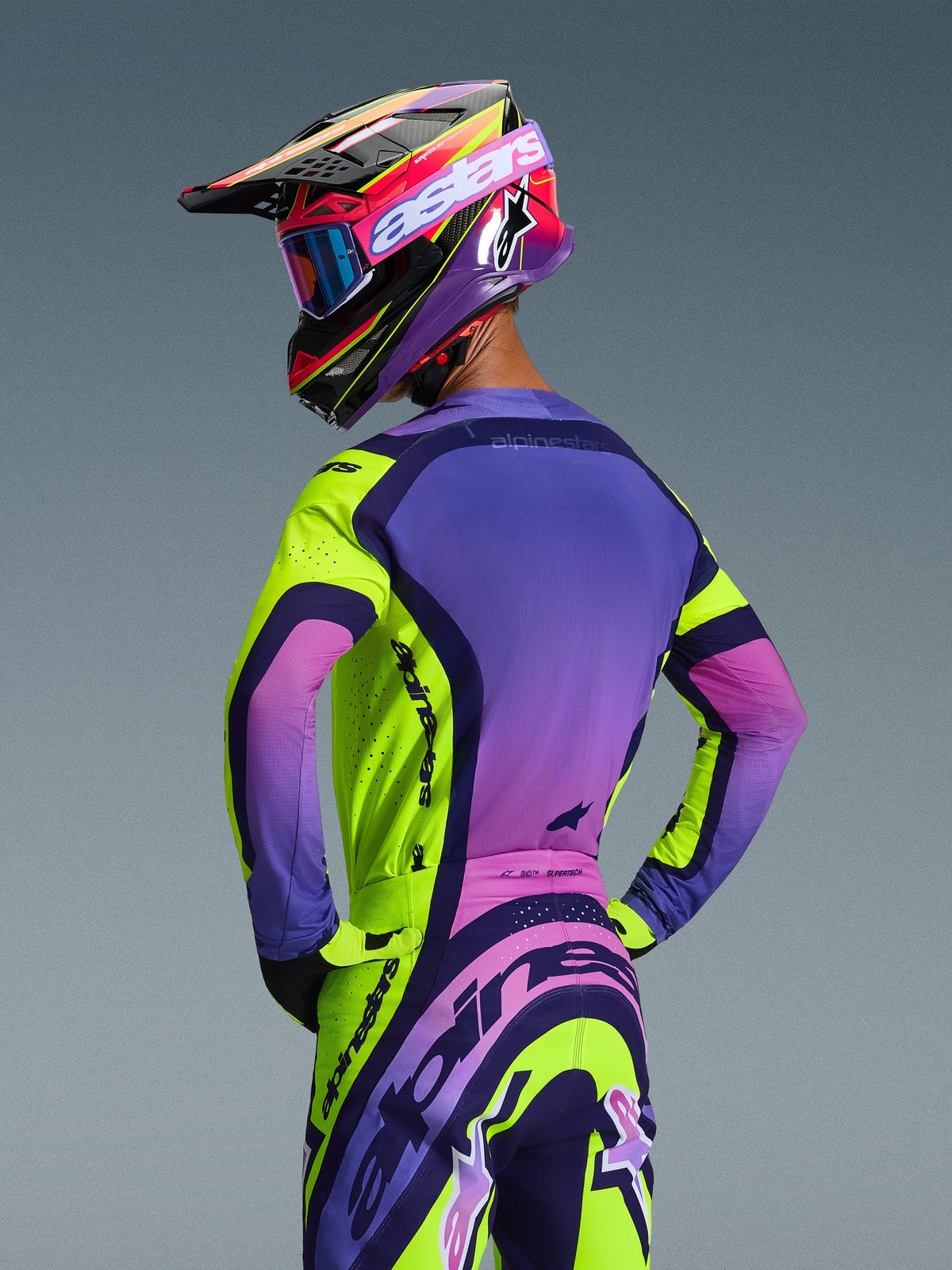 Motocross rider from the back wearing the Alpinestars 2026 Supertech Vista Gear Set, Yellow Purple and Black jersey and pants, featuring a vibrant gradient design, mesh ventilation, and bold Alpinestars branding, coordinated with a purple helmet and goggles.