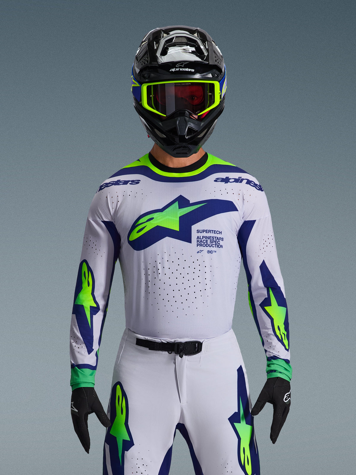 Conjunto de Equipamento Alpinestars 2026 Supertech Vista, camisola e calças de motocross, branco com grafismos em azul e verde néon, usado com capacete preto e óculos néon, com ventilação cortada a laser e ajuste de corrida atlético para competição profissional.