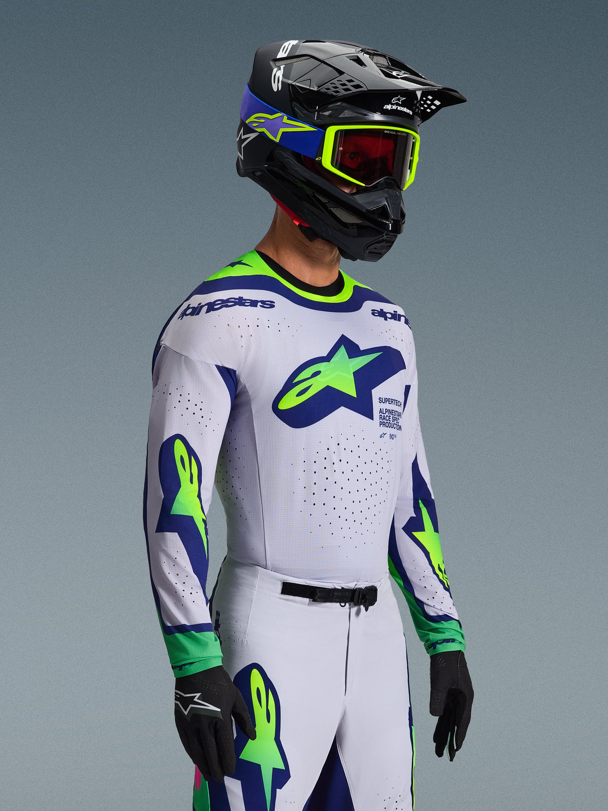 Alpinestars 2026 Supertech Vista Gear Set, camisola e calças de motocross, branco com detalhes em azul e verde-lima, usado por um piloto com capacete preto e óculos de proteção verde néon, com logótipos de estrela proeminentes no peito e nas mangas, corte atlético de nível profissional para performance em todo-o-terreno
