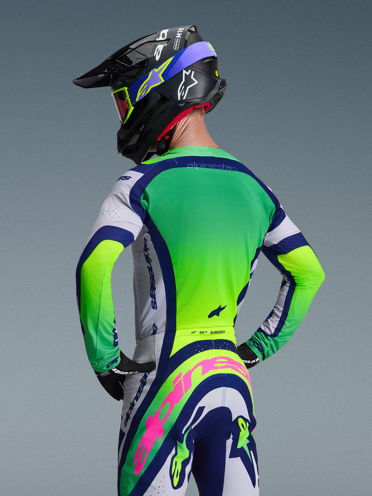 Piloto de motocross visto de costas a usar a camisola Alpinestars 2026 Supertech Vista em Cinzento Claro, Amarelo Fluo e Azul, combinada com calças e capacete a condizer, com painéis néon vibrantes e um corte atlético de corrida para competições off-road.