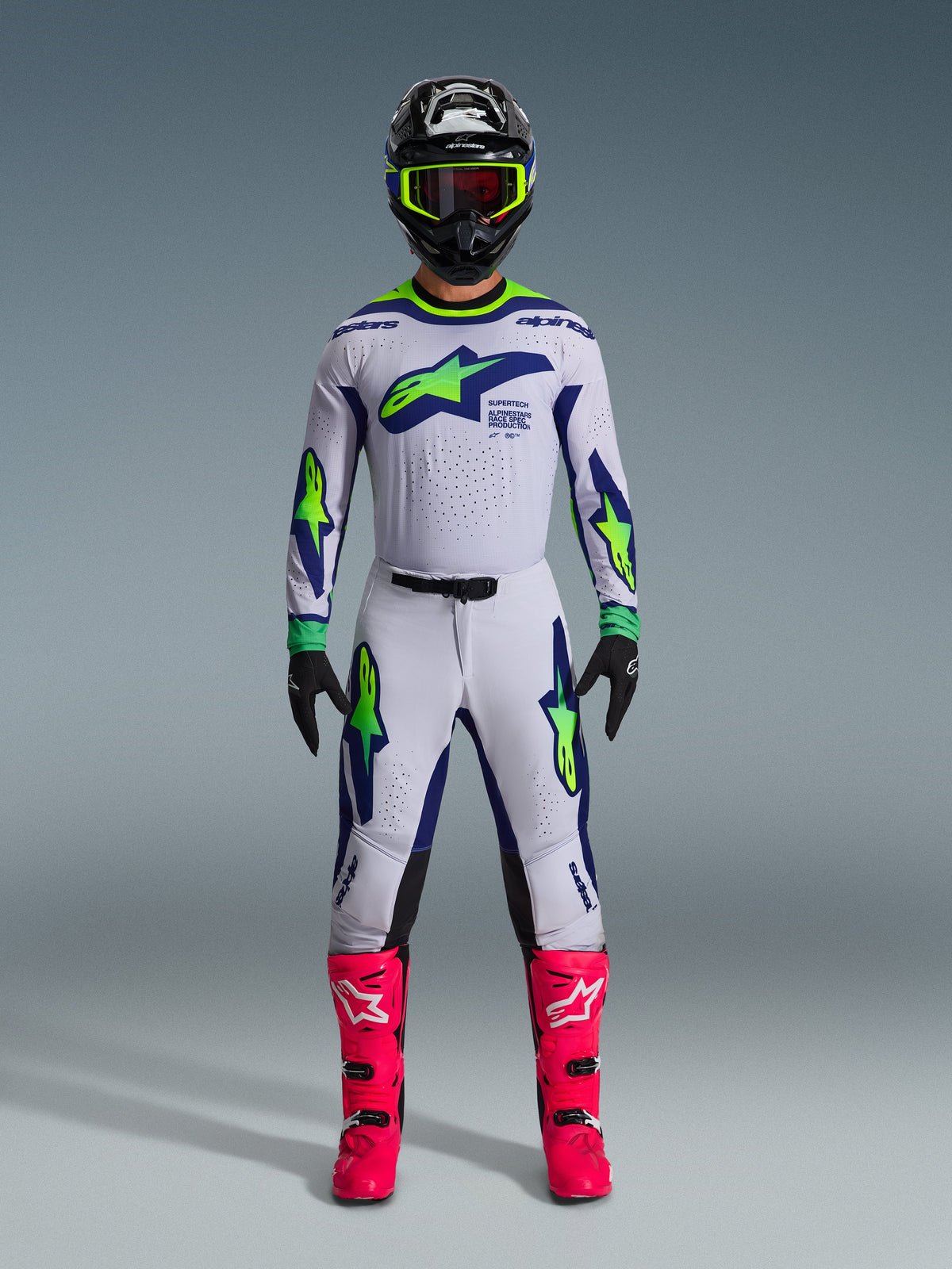 Piloto de motocross a usar a Camisola Supertech Vista 2026 em Cinzento Claro, Amarelo Fluo e Azul, com calças a condizer, botas cor-de-rosa vibrantes e capacete preto com detalhes néon, equipamento de competição off-road de nível profissional