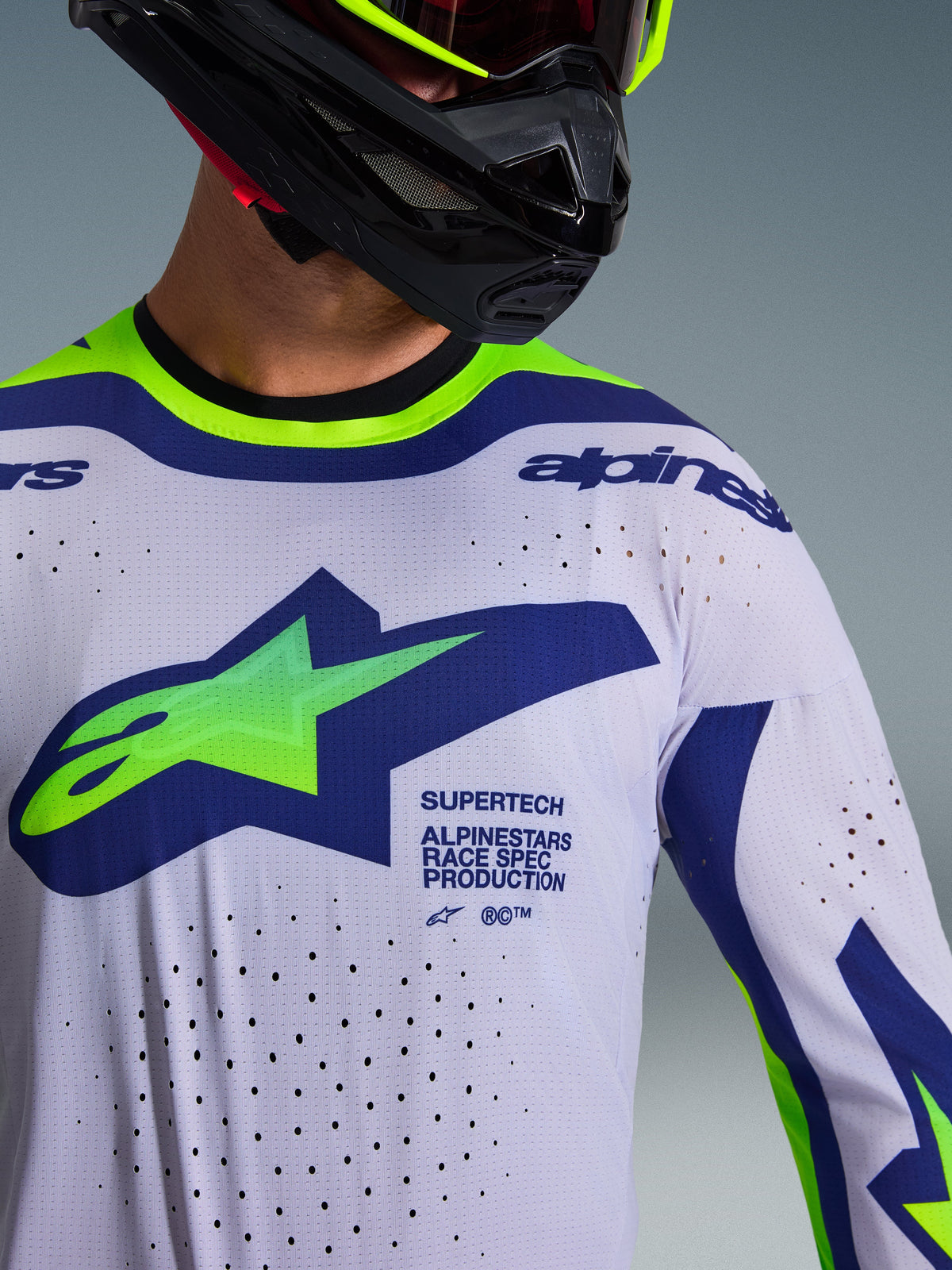 Primeiro plano da camisola Alpinestars 2026 Supertech Vista em cinzento claro, amarelo néon e azul, vestida por um piloto com capacete preto, apresentando um grande logótipo no peito, perfurações de ventilação cortadas a laser e branding profissional de competição.