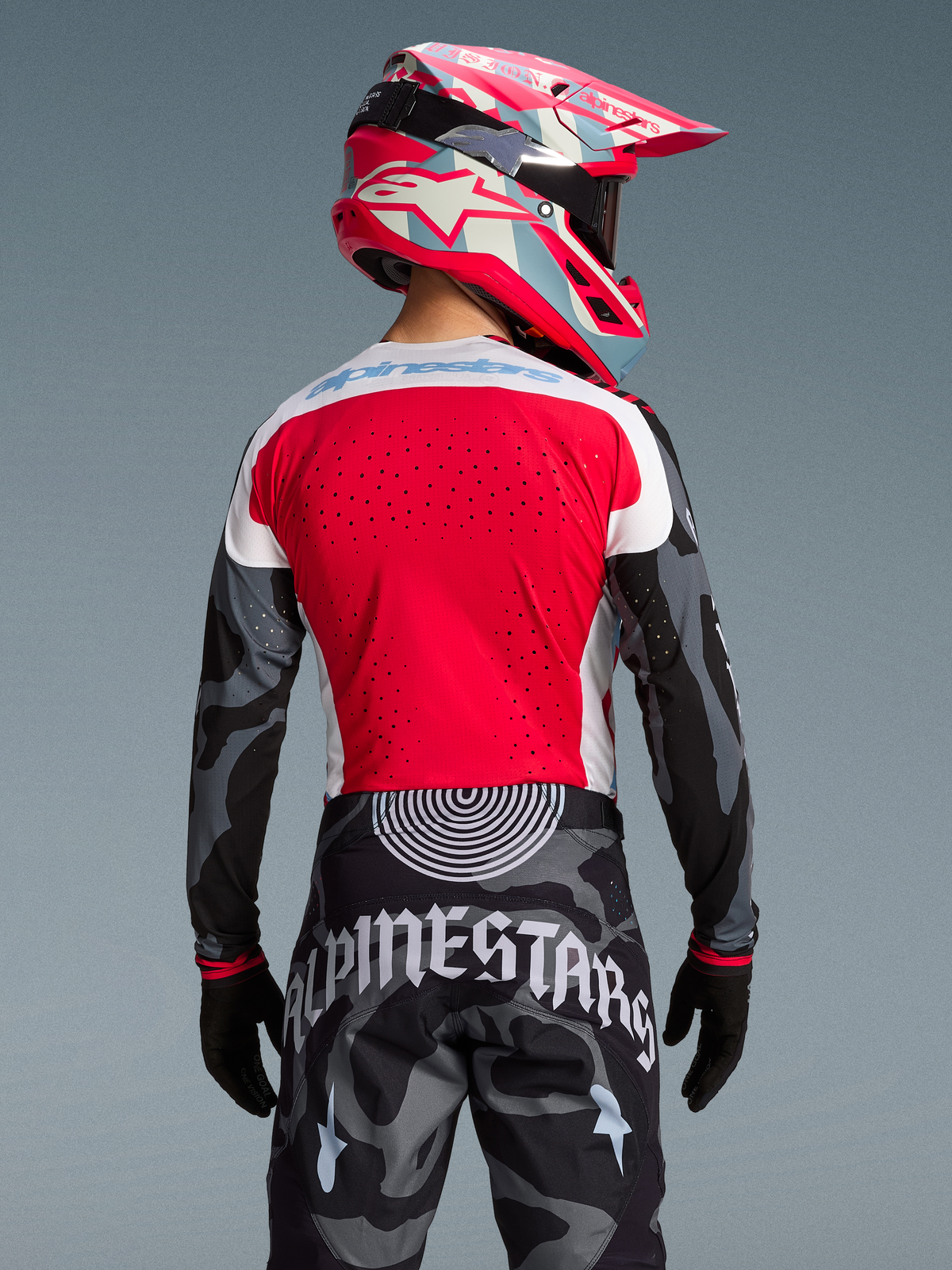 Camisola Alpinestars Techstar Beast Coast, de costas com camisola vermelha perfurada, calças camufladas e capacete rosa.