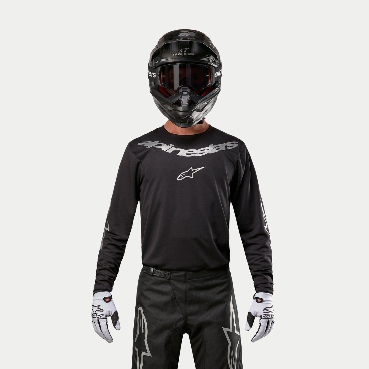 Alpinestars 2024 Fluid Graphite Jersey, camisola de motocross em preto e prateado, usada por um piloto com capacete preto e óculos vermelhos, com logótipo Alpinestars prateado no peito, combinada com calças pretas e luvas brancas para performance todo-o-terreno