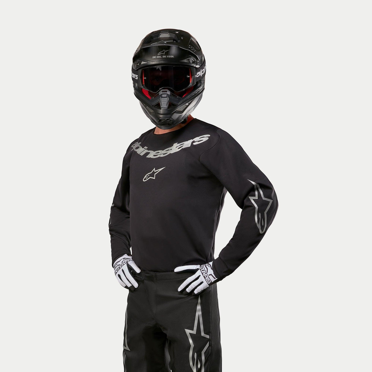 Piloto de motocross a usar a camisola Alpinestars 2024 Fluid Graphite, em preto e prateado, combinada com calças de motocross pretas, capacete preto com óculos de lentes vermelhas e luvas brancas e pretas, com logótipos prateados da marca no peito e nas mangas
