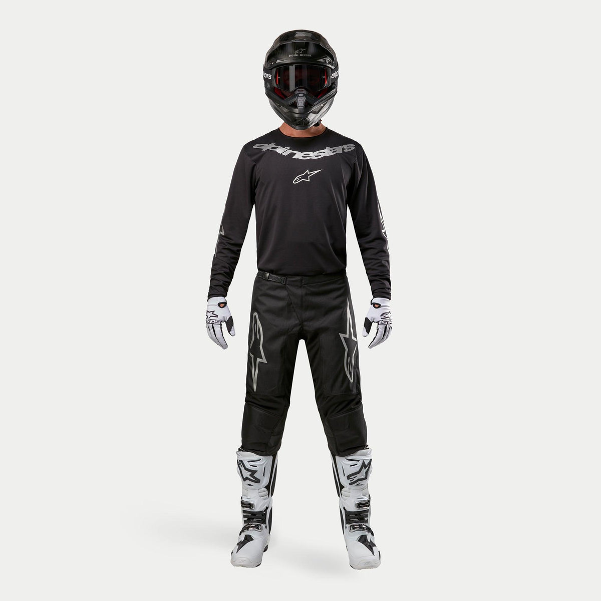 Piloto de motocross com equipamento de corrida completo, Camisola Alpinestars 2024 Fluid Graphite em preto e prata, calças pretas com logótipos de estrela prateados, capacete preto com óculos vermelhos, botas brancas e pretas, luvas brancas e pretas, conjunto de motocross coordenado