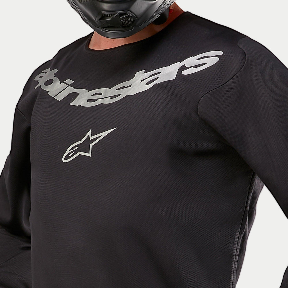 Alpinestars 2024 Fluid Graphite Jersey, camisola de motocross, preta e prateada, detalhe em grande plano da zona do peito com a marca Alpinestars e logótipo de estrela prateados, construção em poli-tecido respirável, usada por um piloto com capacete preto