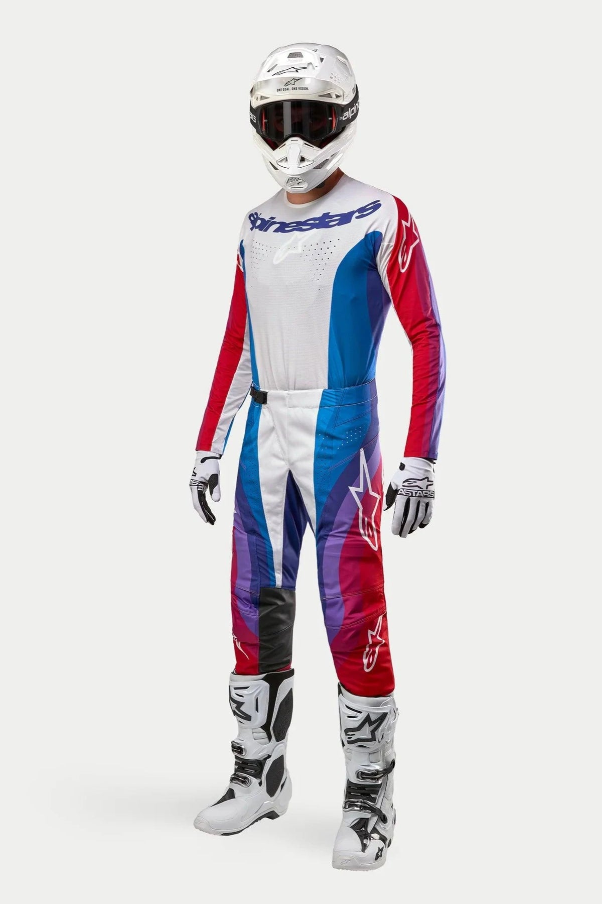 Piloto de motocross com equipamento completo Alpinestars, calças Techstar Pneuma 2024 em azul, vermelho e branco, camisola de manga comprida a condizer, capacete branco com óculos escuros e botas brancas e pretas, conjunto coordenado para corridas off-road.