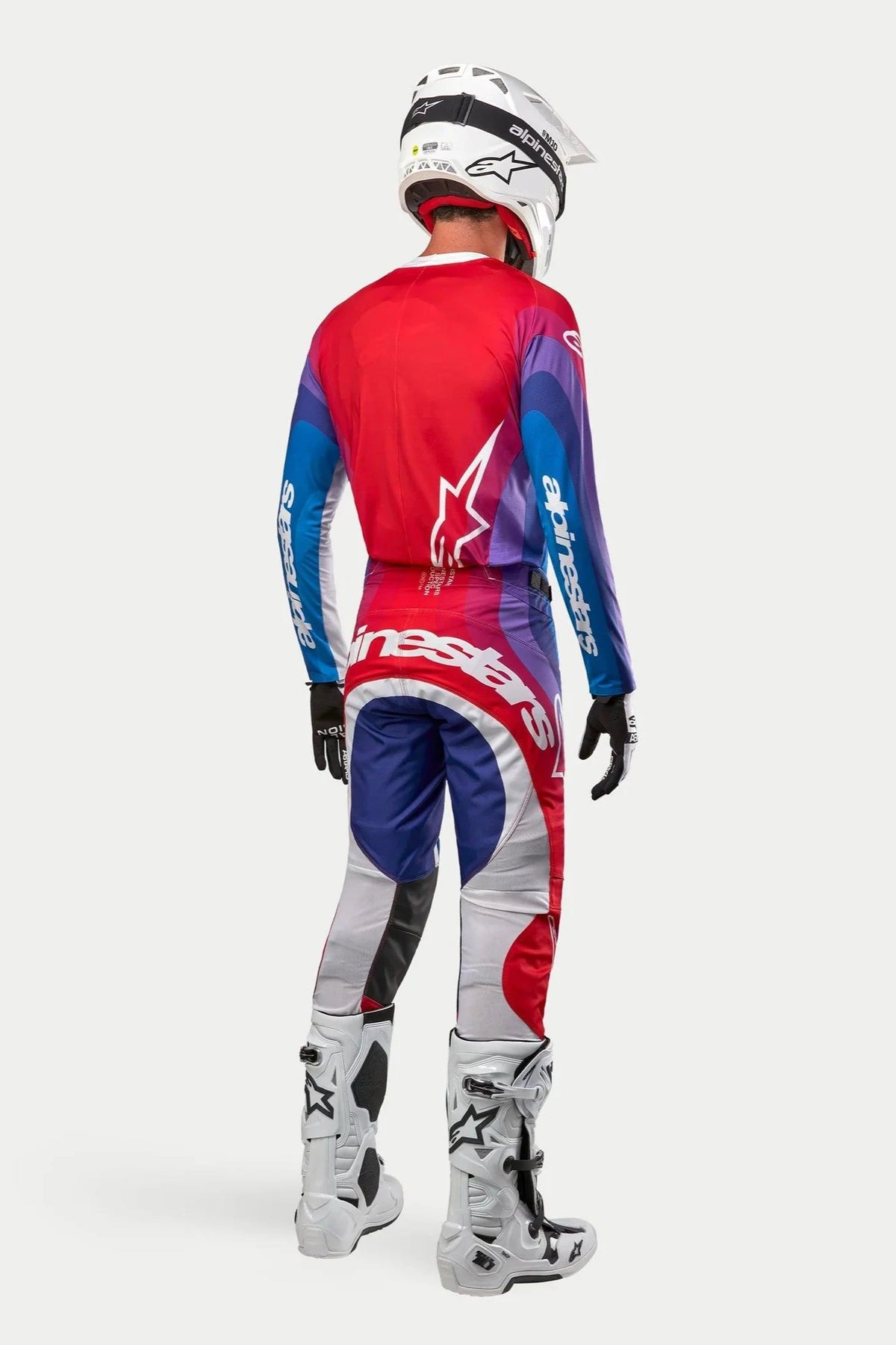 Piloto de motocross visto de trás com camisola e calças Alpinestars 2024 Techstar Pneuma, esquema de cores Blu Mars Red White com gradientes de vermelho, azul e roxo, logótipos brancos, botas brancas e capacete branco, equipamento de corrida off-road coordenado.