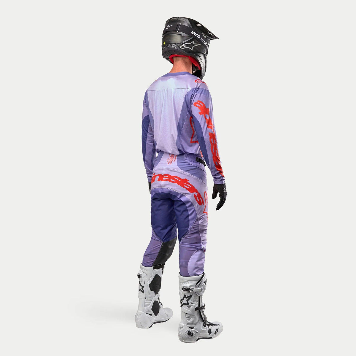 Equipamento completo de motocross Alpinestars 2024 Techstar Ocuri, com camisola e calças em tons de roxo e laranja, vista traseira do piloto, incluindo capacete preto, luvas pretas e botas brancas com detalhes pretos