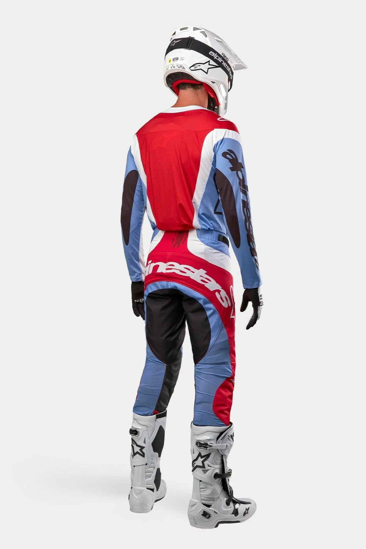 Motocross rider wearing complete racing gear from behind, 2024 Techstar Ocuri Jersey and Pants in Light Blue, Mars Red, and White, white Alpinestars helmet with black graphics, white and grey off-road boots, black gloves, coordinated outfit for motocross racing
