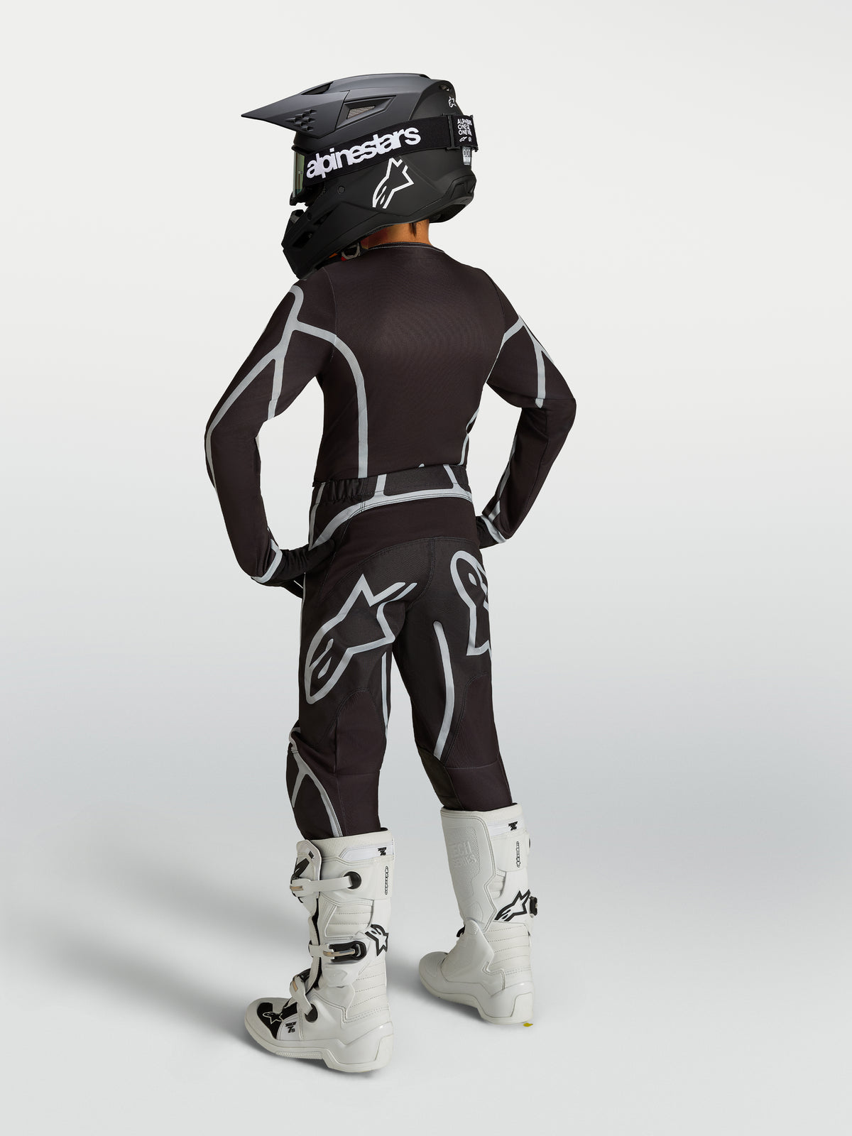 Conjunto Alpinestars 2027 Juventude Fluid Apex V2 de motocross, preto e prateado, vista traseira com botas brancas.