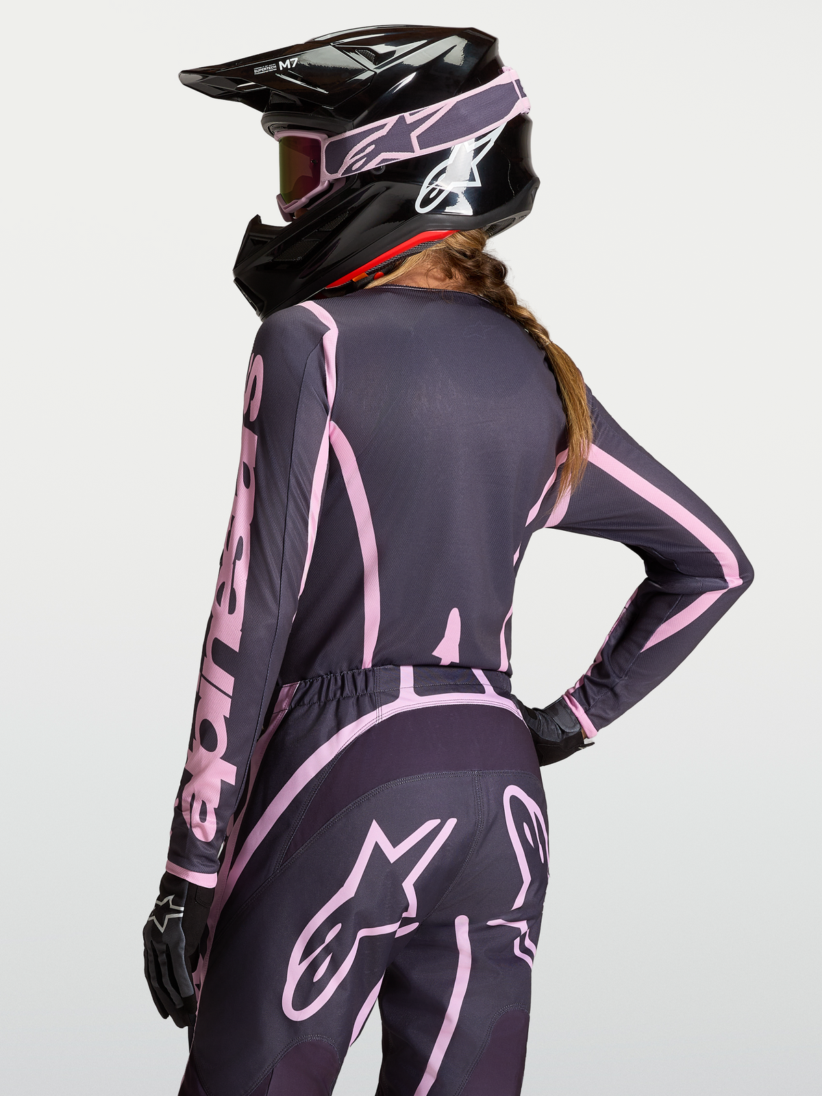 Alpinestars Camisola Stella Fluid Apex V2, equipamento de motocross para mulher, cinzenta e rosa, vista traseira on-model com calças a condizer e capacete preto.